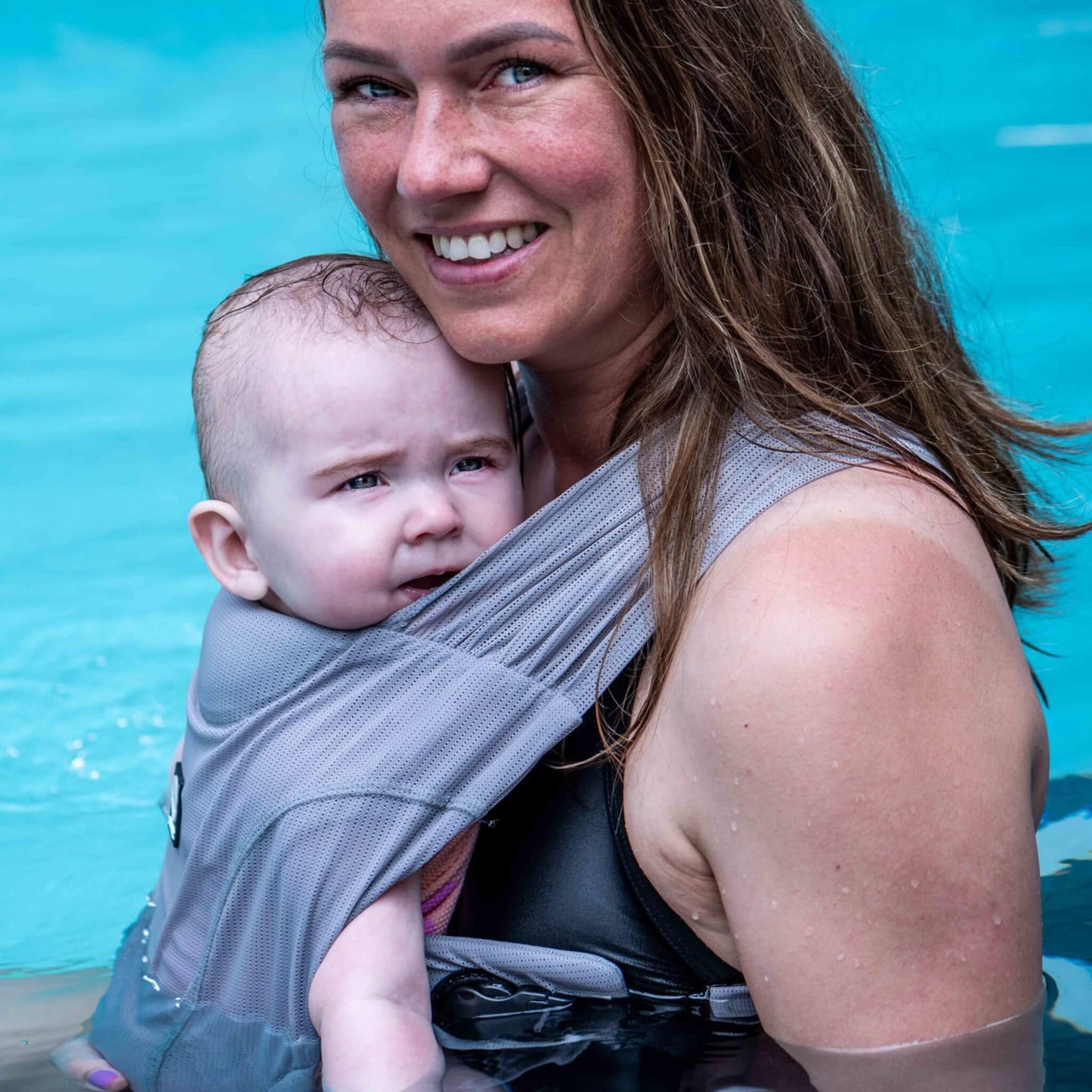 A parent standing in a swimming pool with a baby held against their chest in a grey water baby carrier.