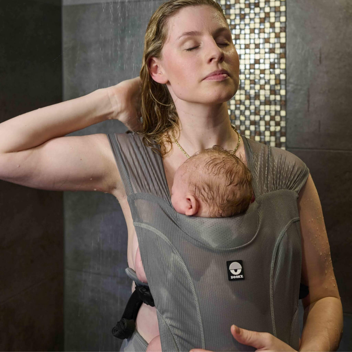 A woman showering while carrying a young baby close against their chest in a grey water baby carrier.