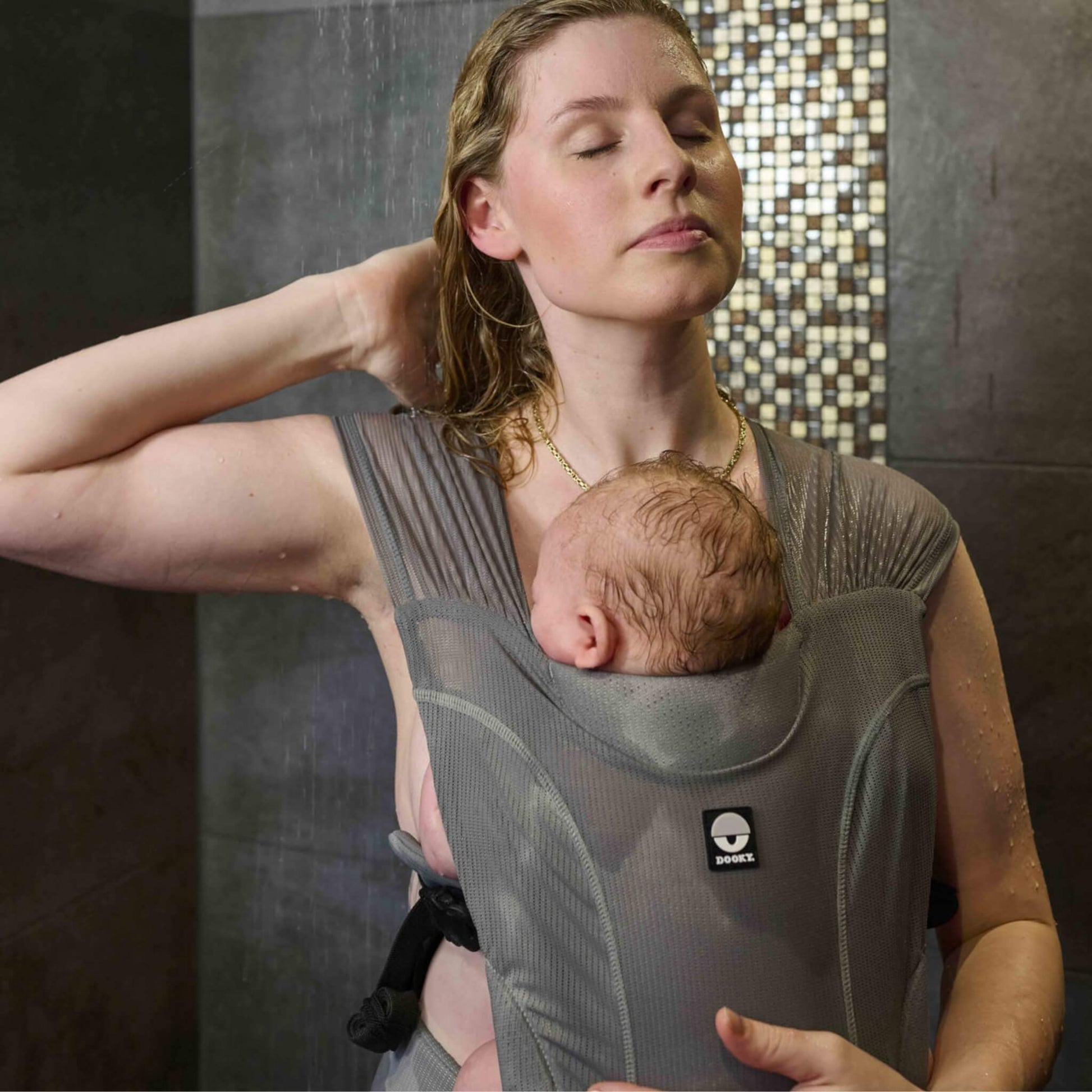 A woman showering while carrying a young baby close against their chest in a grey water baby carrier.