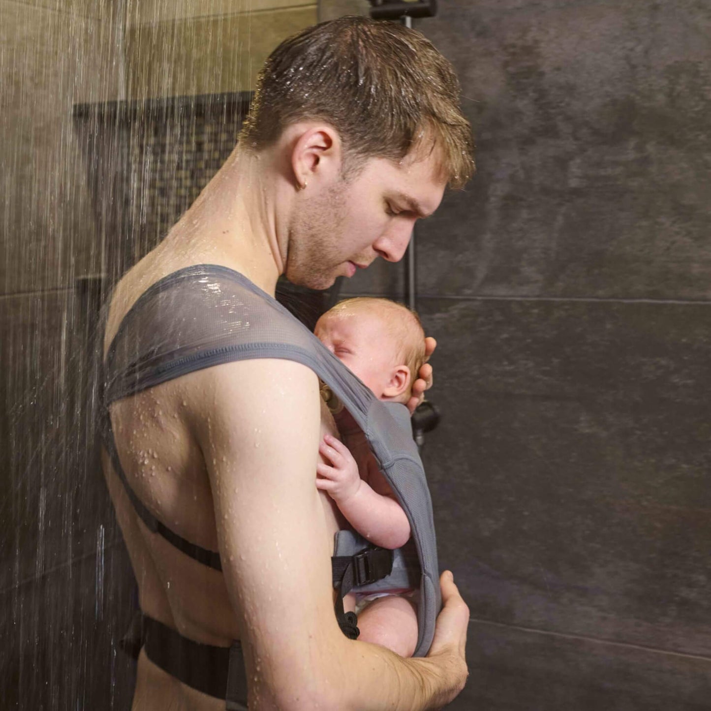 A parent holding a young baby securely against their chest in a grey baby carrier while showering.