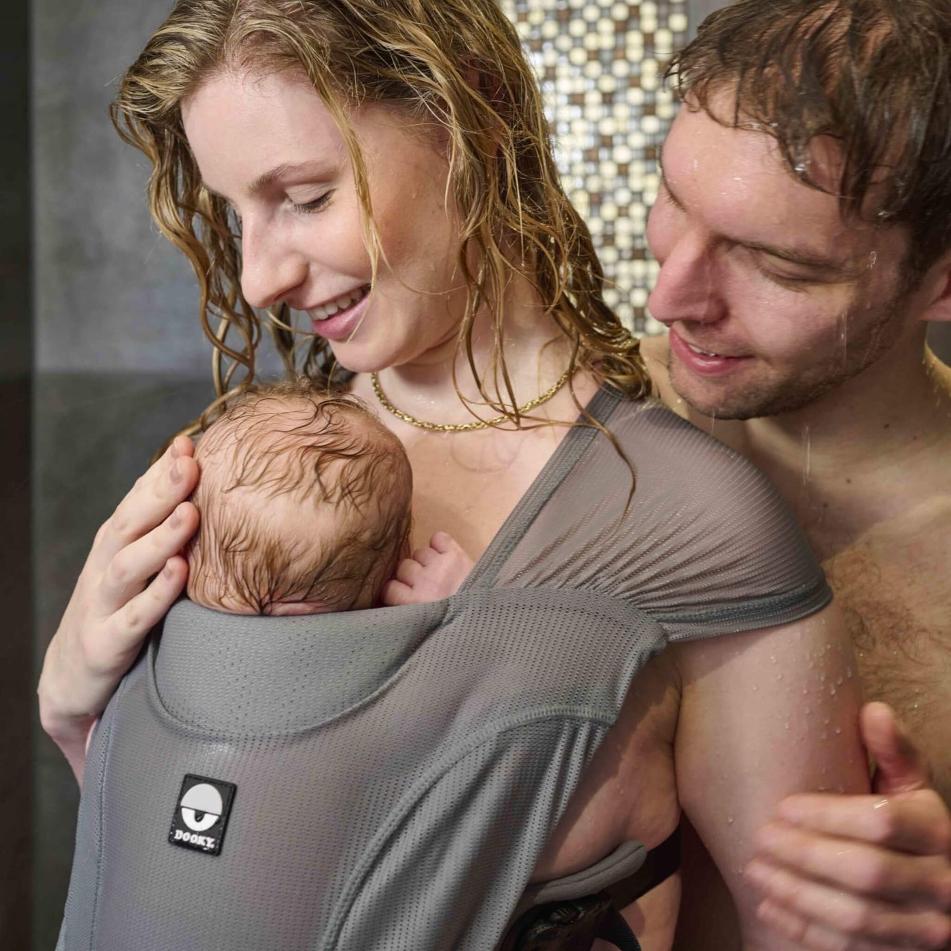 A woman and a man holding a baby in a gray mesh baby wrap carrier, photographed in a real indoor setting with water on their hair and skin.