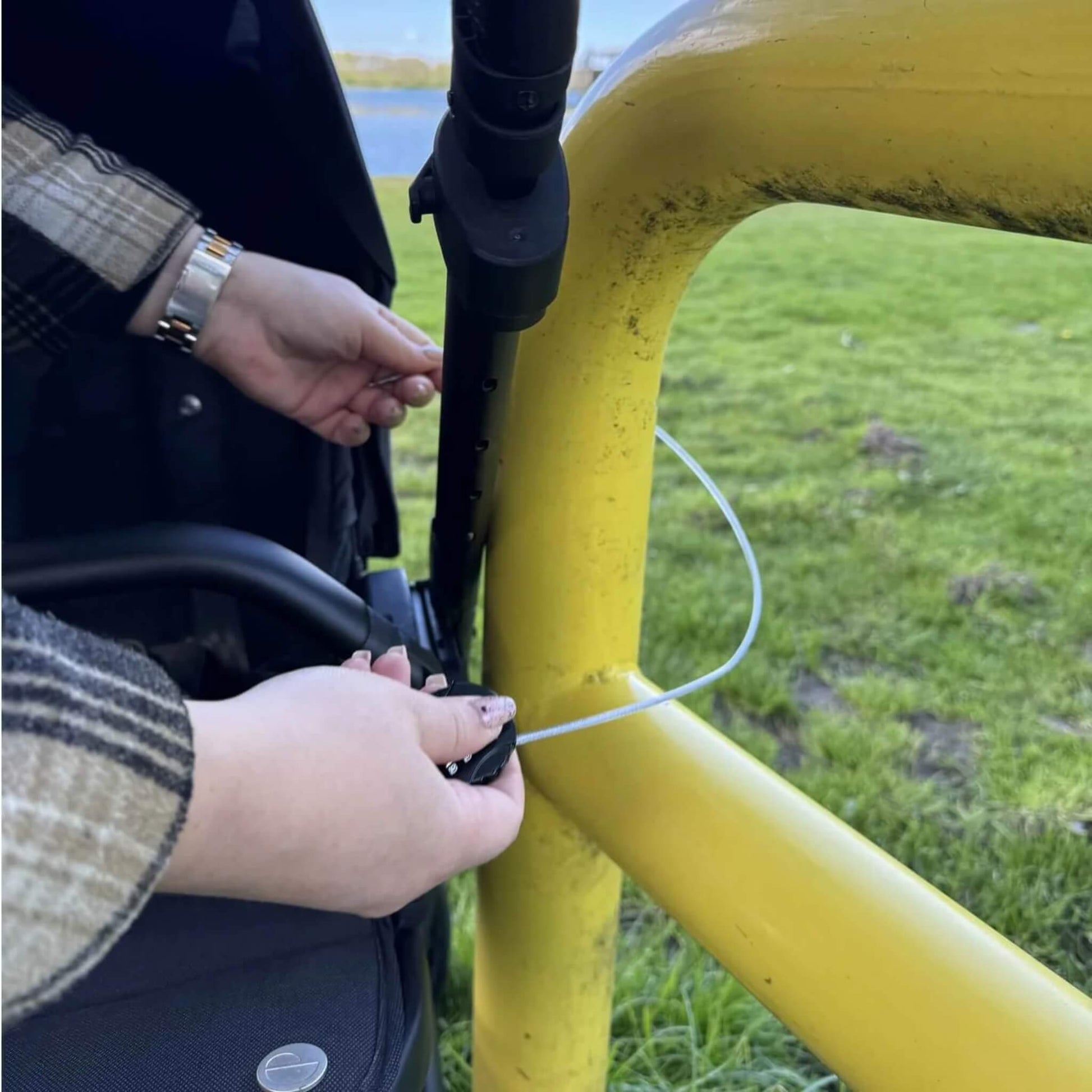 A person threading a silver cable lock through a stroller frame and around a yellow metal barrier outdoors.