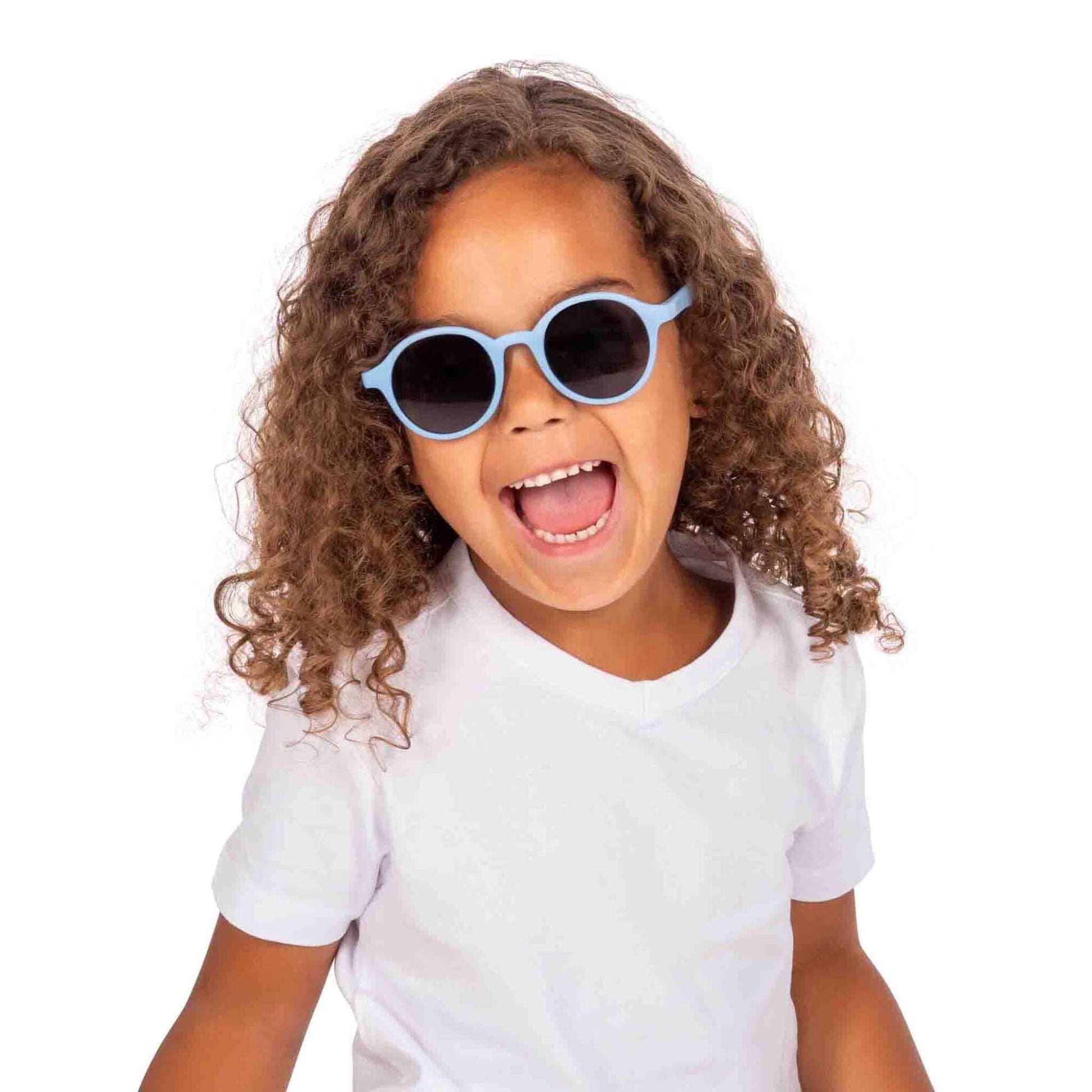 A young child with curly hair wearing blue round sunglasses and a white t-shirt, smiling brightly against a plain background.