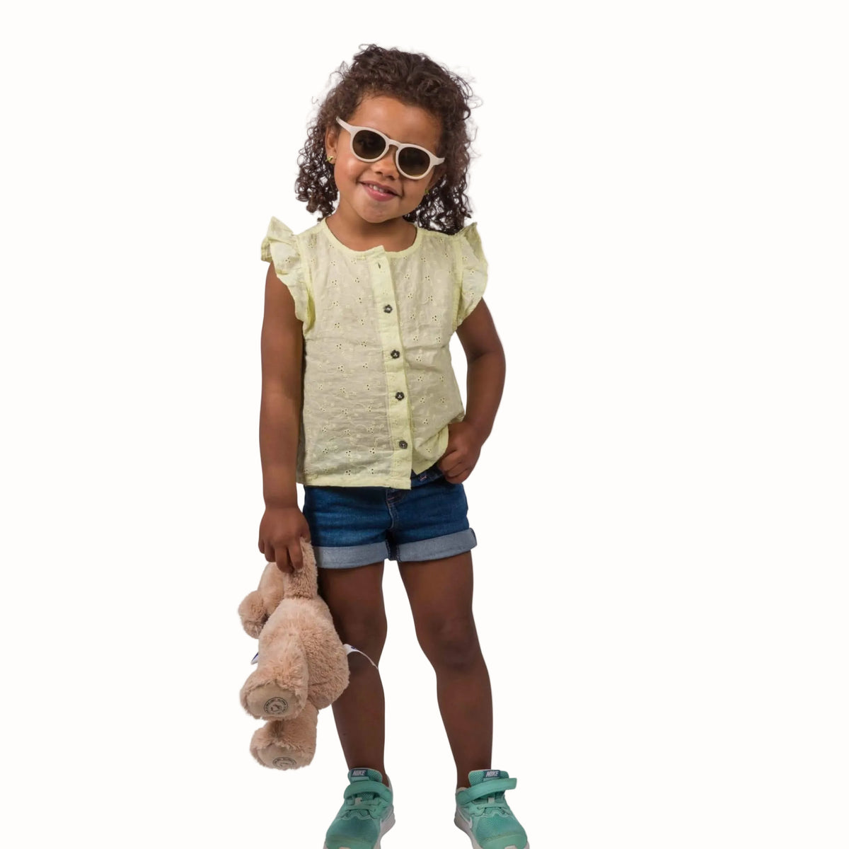 A young girl with curly brown hair wearing round white-framed sunglasses, a pale yellow ruffle-sleeve button-front top, denim shorts, and mint green sneakers, holding a light brown plush toy against a solid white background.