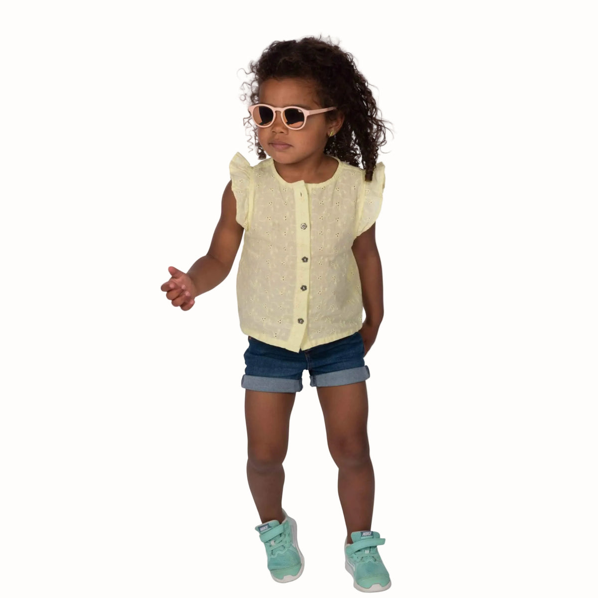 A toddler girl with curly hair wearing a pale yellow sleeveless button-front top, denim shorts, and mint green sneakers, standing against a plain white background.