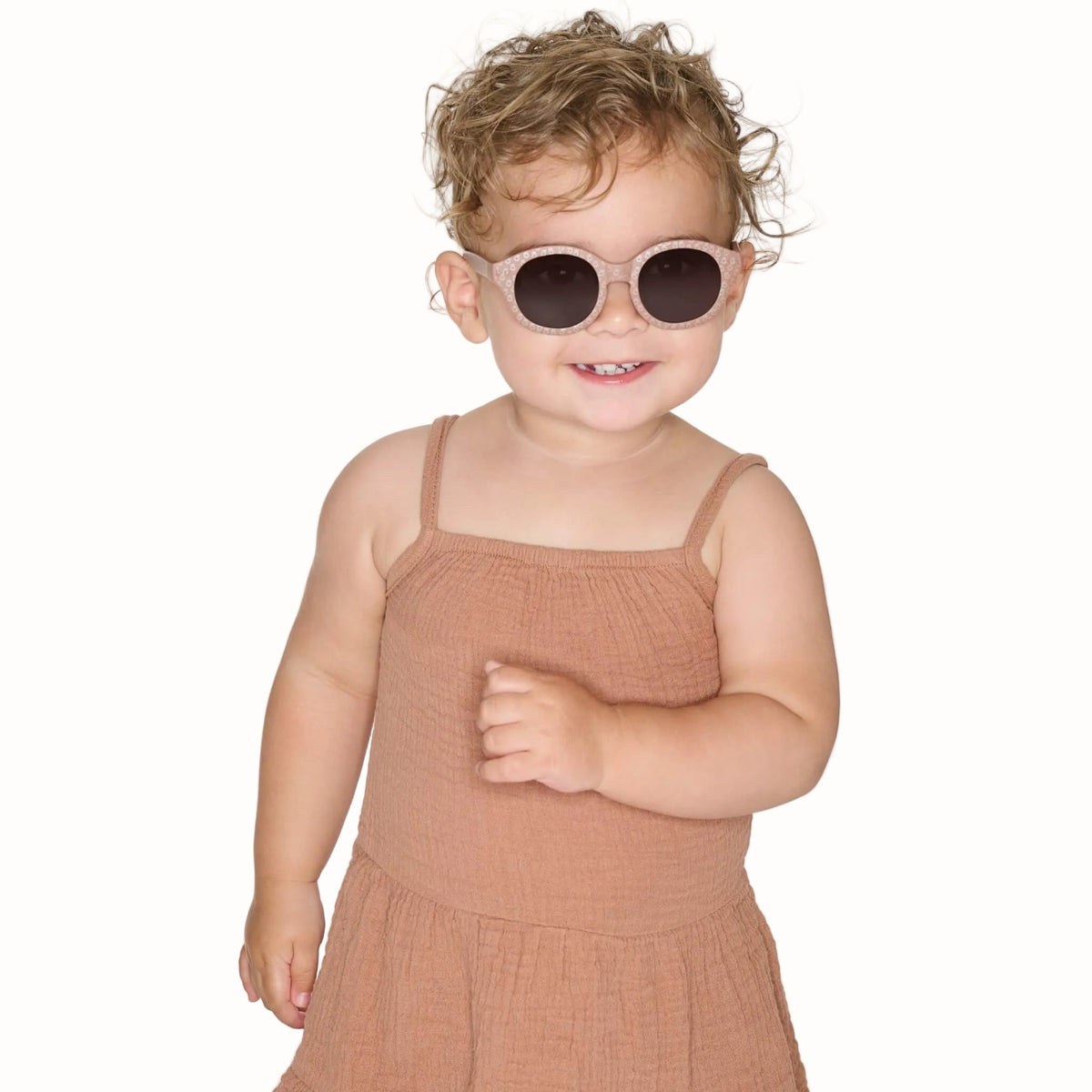 Toddler wearing taupe patterned sunglasses with rounded dark lenses and a brown sleeveless outfit.