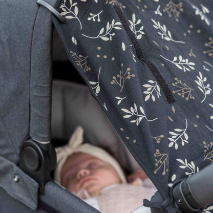 A side angle of a sleeping baby inside a pram beneath the softly draped black sunshade in a romantic leaf design, partially shading the infant’s face.