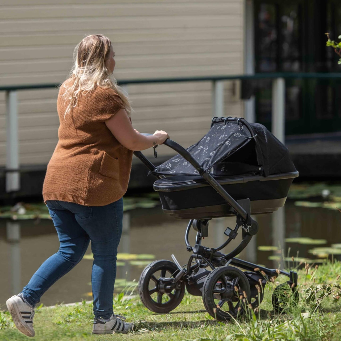 A woman in jeans and a brown top pushes a stroller outdoors, with a black leaf-patterned sunshade covering the pram’s carrycot as she walks past a pond.