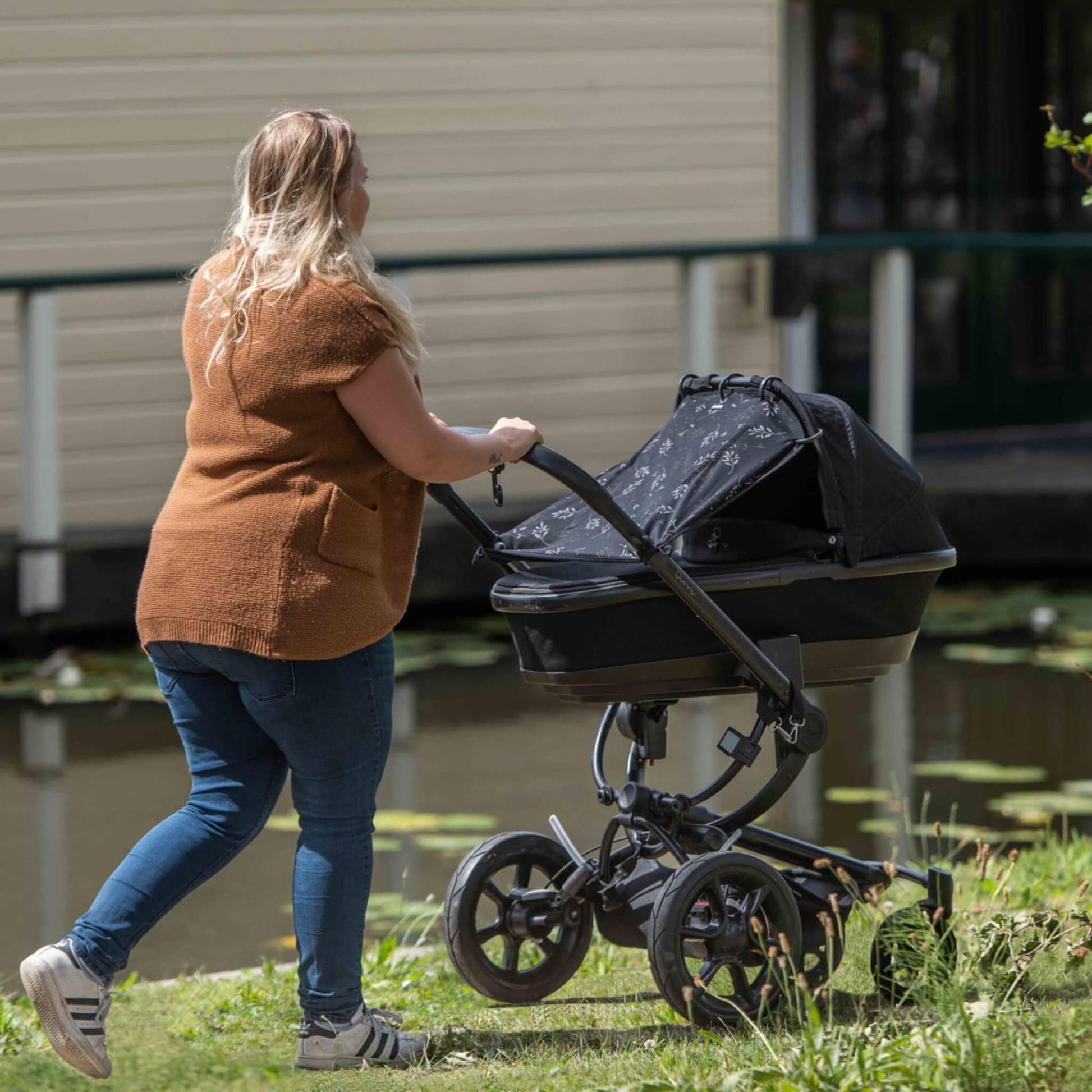A woman in jeans and a brown top pushes a stroller outdoors, with a black leaf-patterned sunshade covering the pram’s carrycot as she walks past a pond.