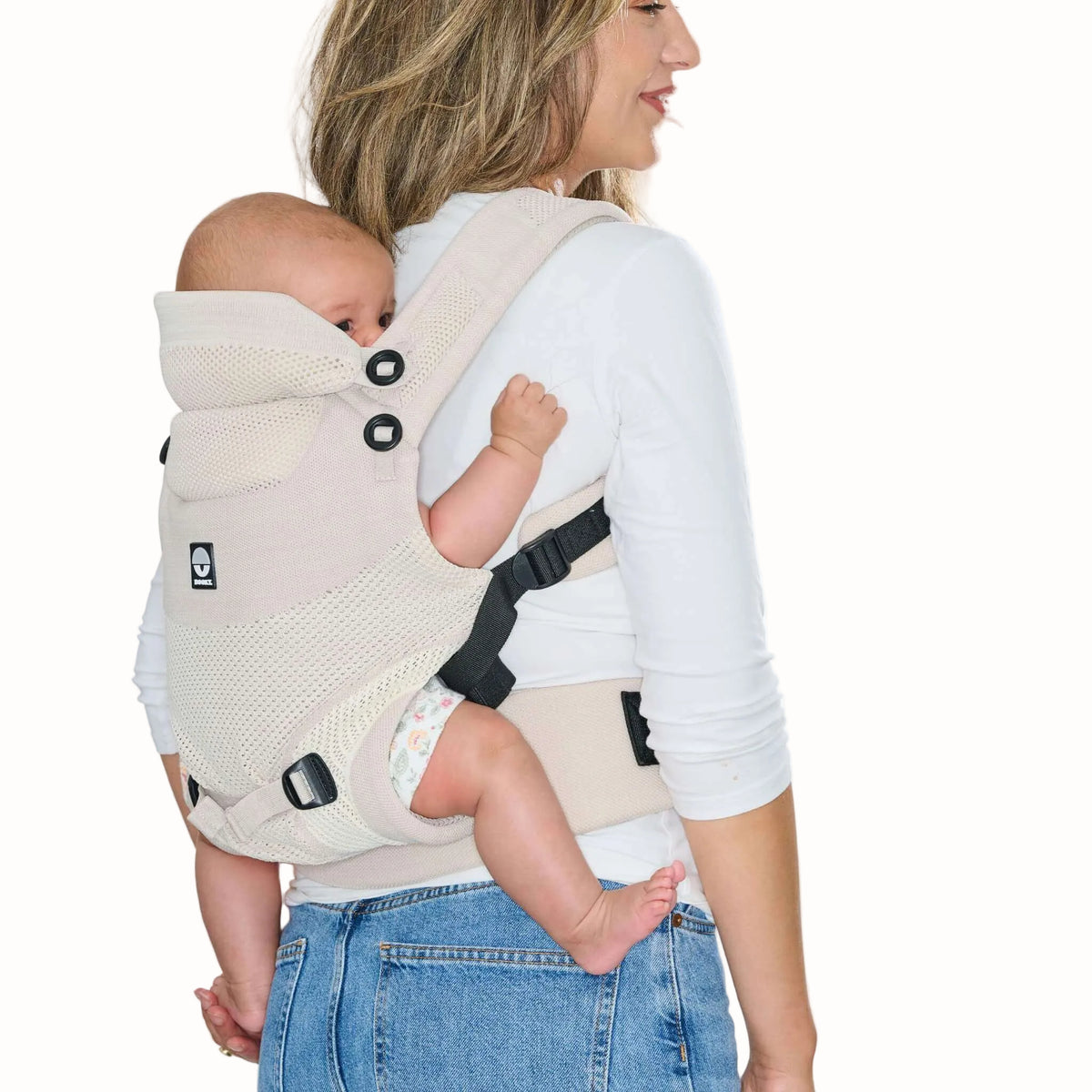 A baby carried on an adult's back in a beige baby carrier, showing the supportive seat, head support and adjustable straps.