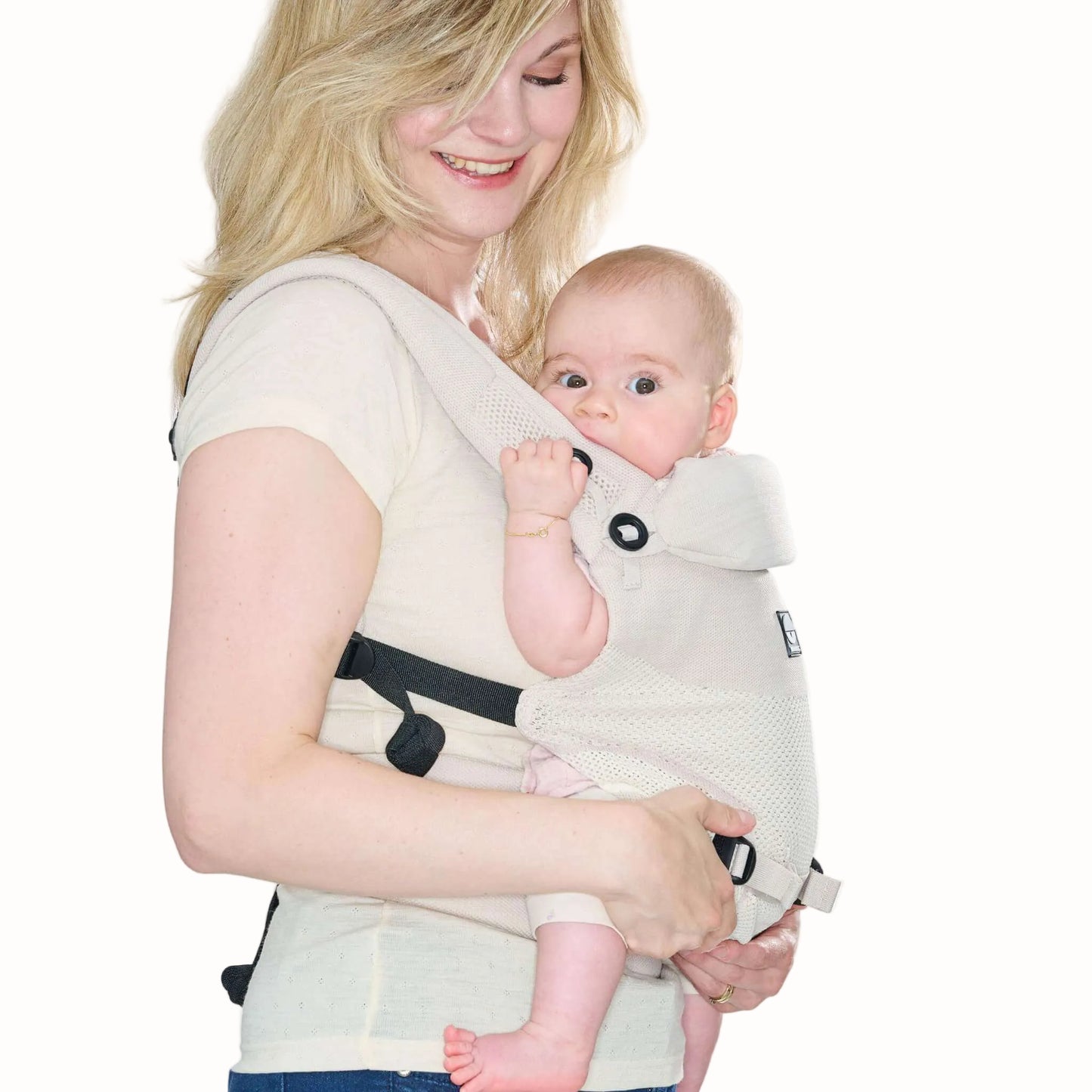 A baby carried facing inwards in a beige baby carrier, with the padded head support folded up across the front.