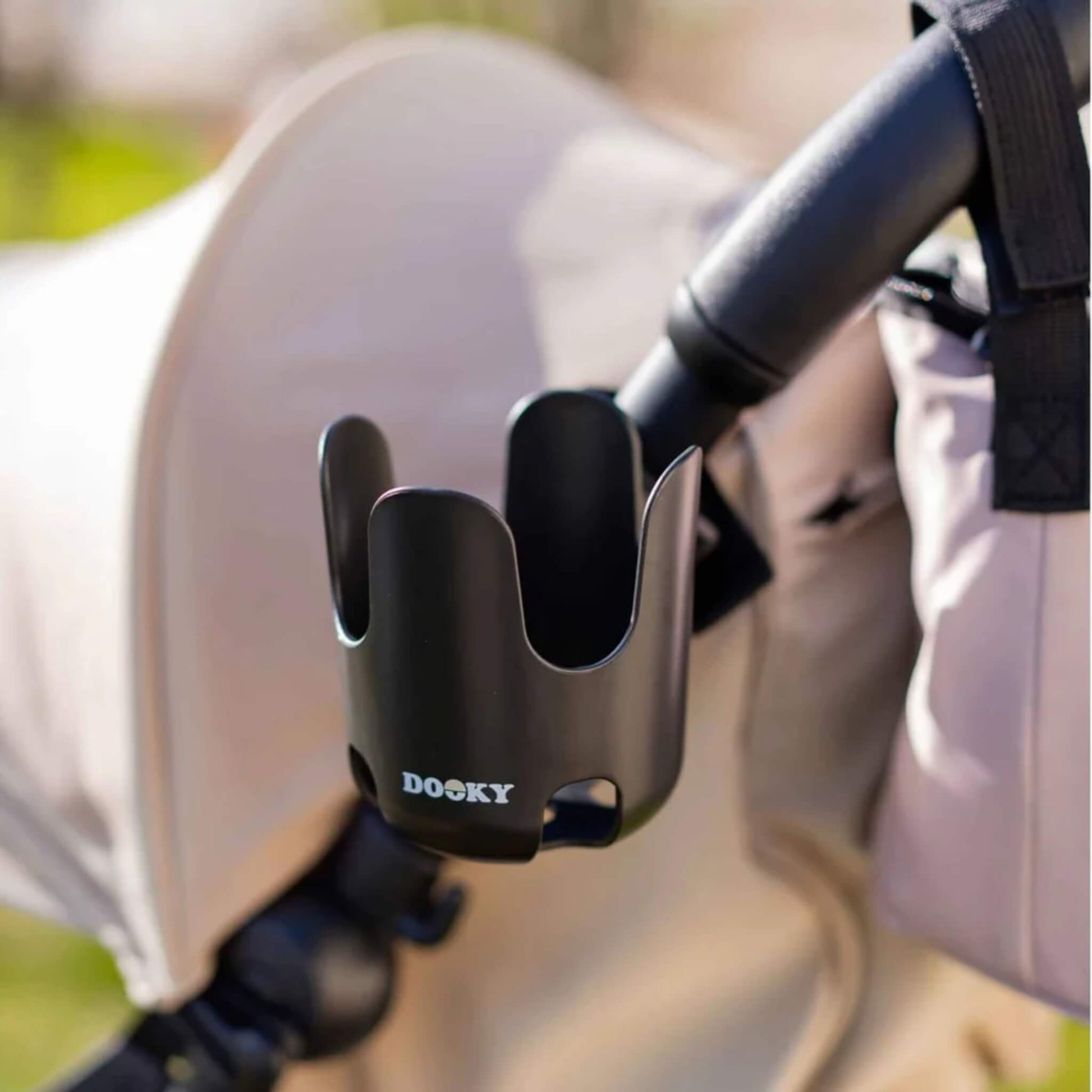 A black cup holder mounted on the side of a stroller handle with a beige canopy and a bag attached nearby.