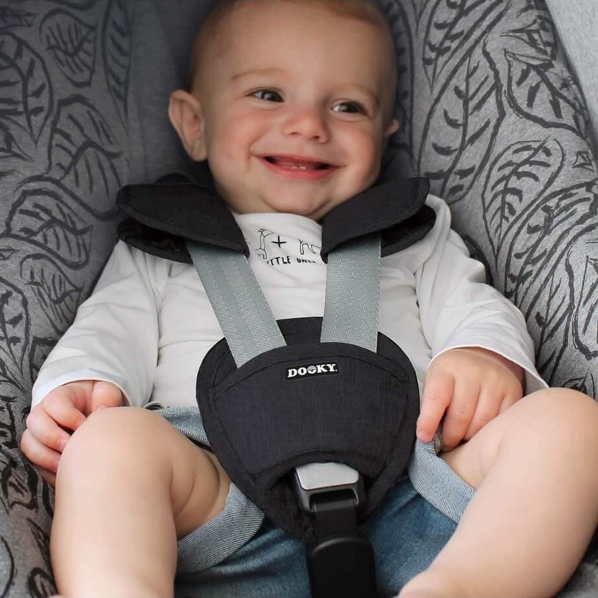 Smiling baby seated in a patterned car seat, wearing a harness fitted with black padded strap covers and buckle pad.