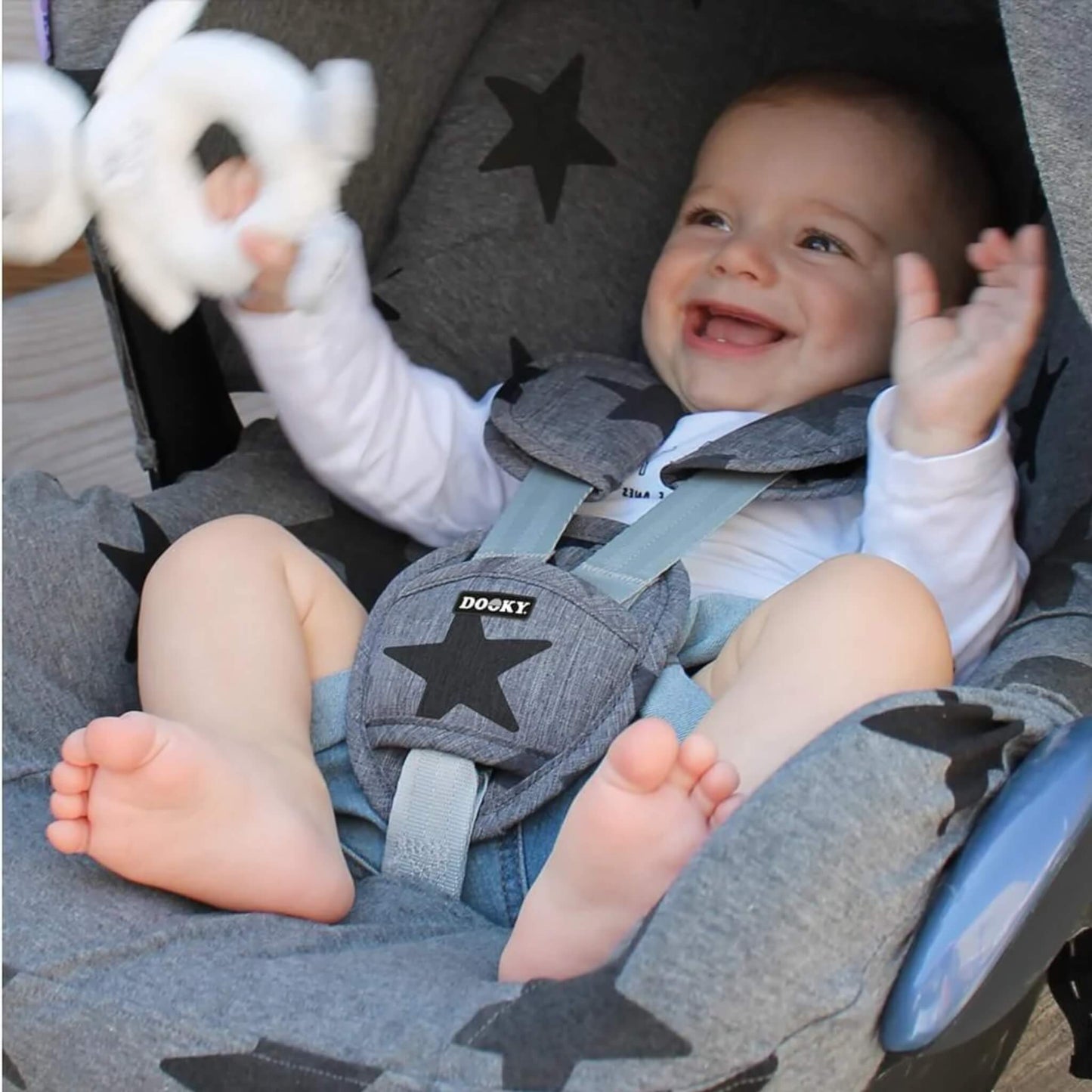Baby smiling and holding a toy while seated in a car seat, wearing a harness fitted with grey star-patterned padded strap covers and buckle pad.