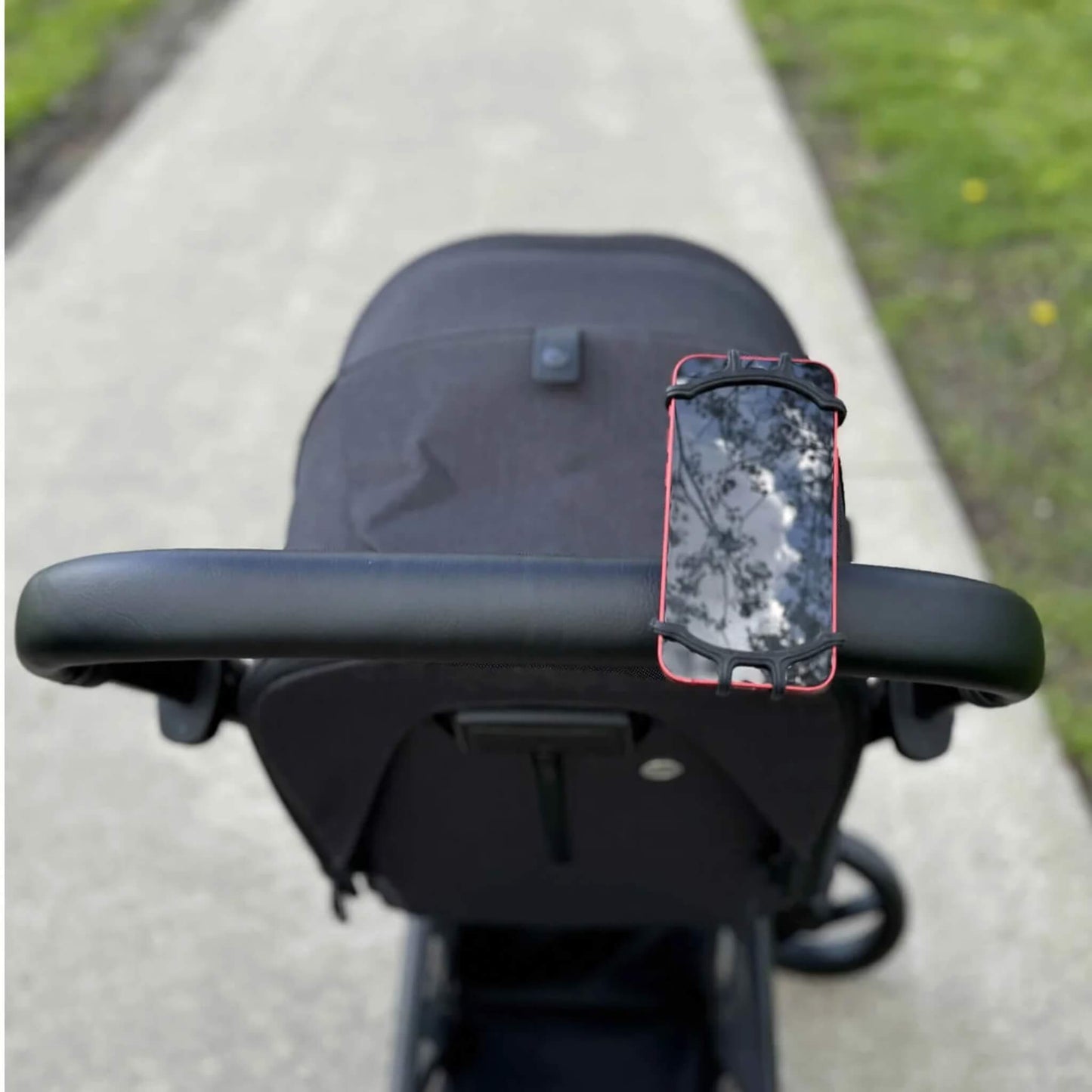 A black phone holder mounted on a stroller handlebar holding a red smartphone outdoors on a paved path.