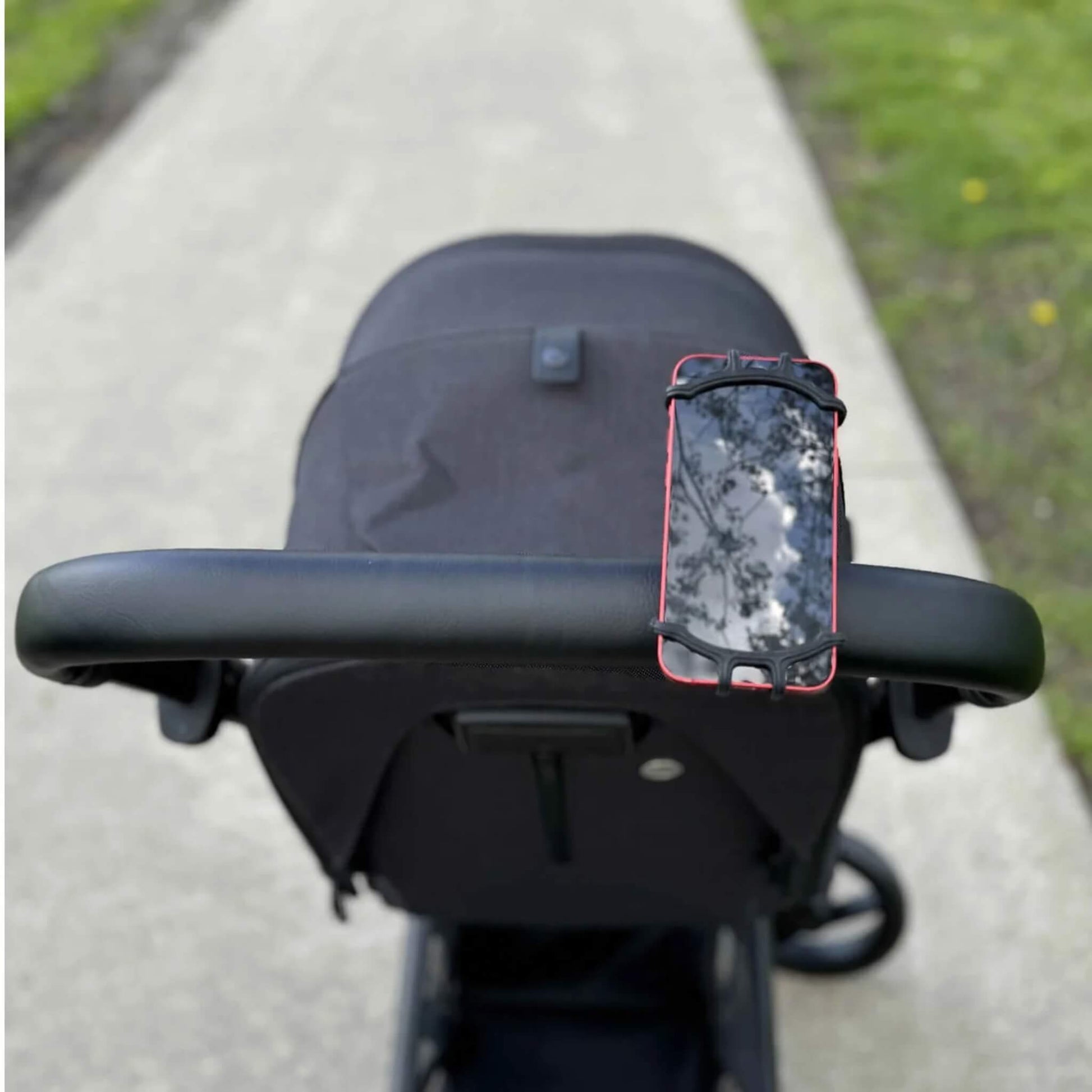 A black phone holder mounted on a stroller handlebar holding a red smartphone outdoors on a paved path.