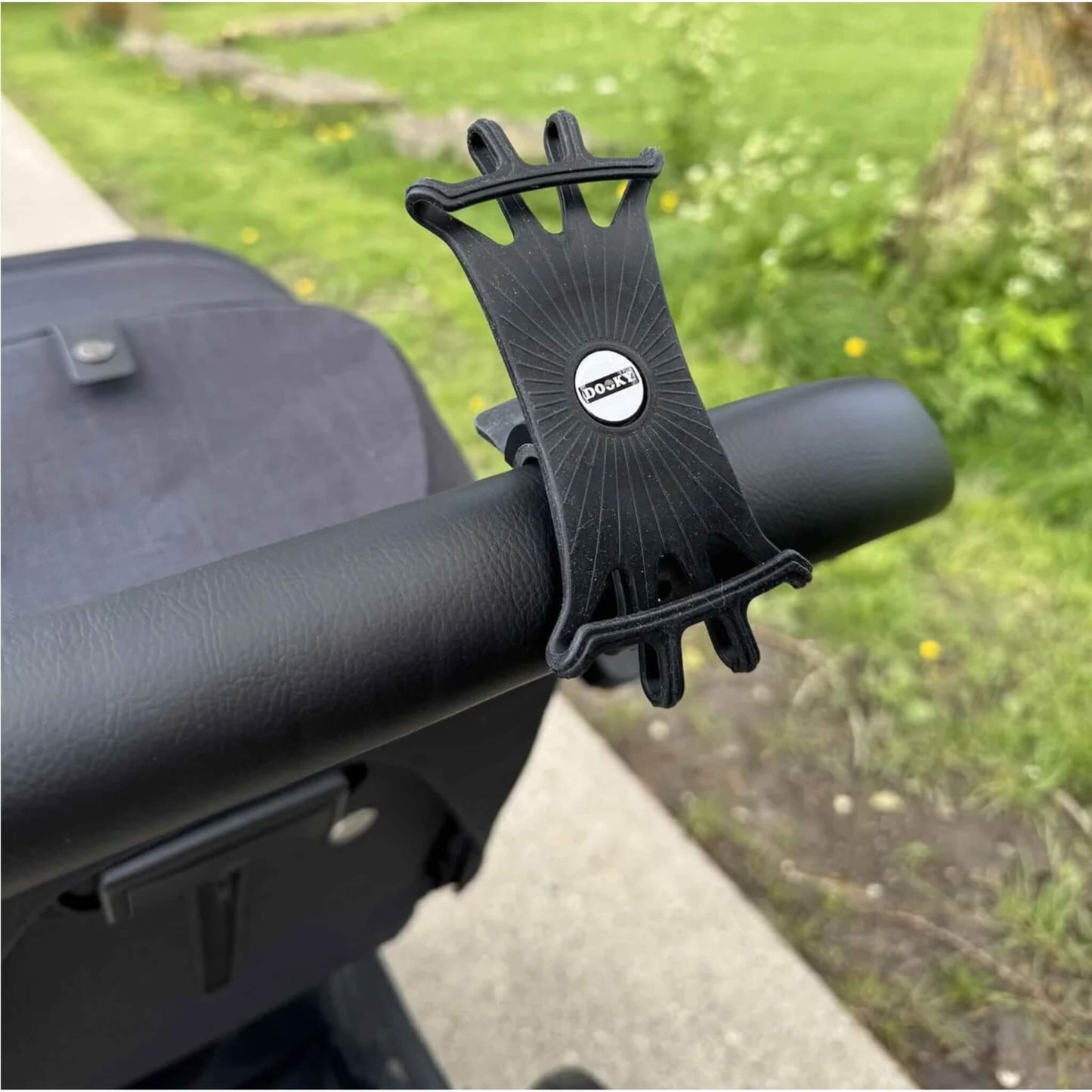 A flexible black phone holder attached to a black stroller handlebar with grass in the background.