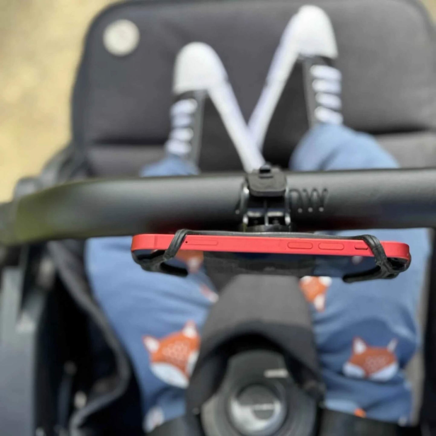 A red smartphone held horizontally in a phone holder on a stroller handlebar, with a child’s legs in fox-print trousers visible below.