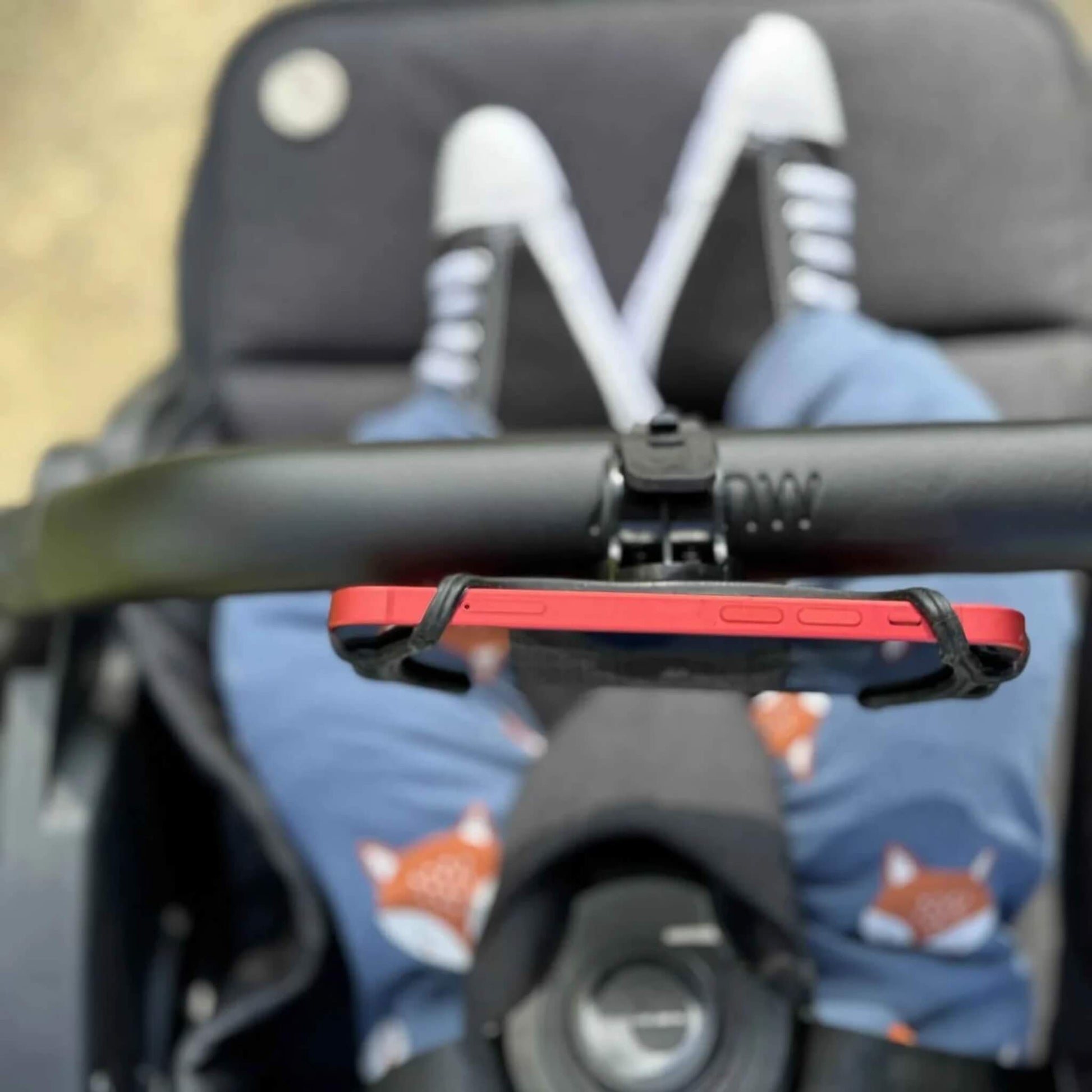 A red smartphone held horizontally in a phone holder on a stroller handlebar, with a child’s legs in fox-print trousers visible below.