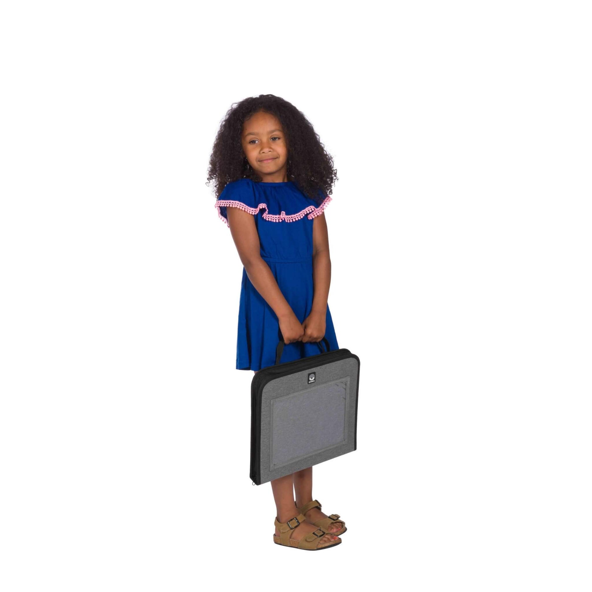 A young girl wearing a blue dress and brown sandals stands upright, holding a closed grey travel tray by the carry handle with both hands.