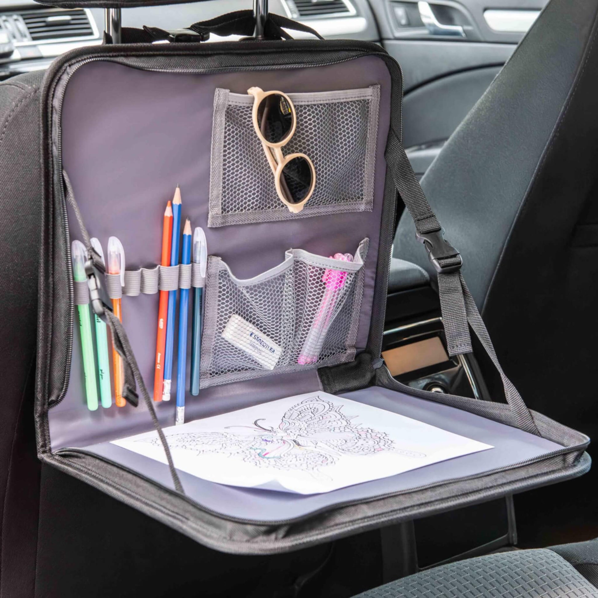A car back seat organiser filled with art supplies including coloured pencils, pens and sunglasses, with a butterfly colouring page resting on the tray.