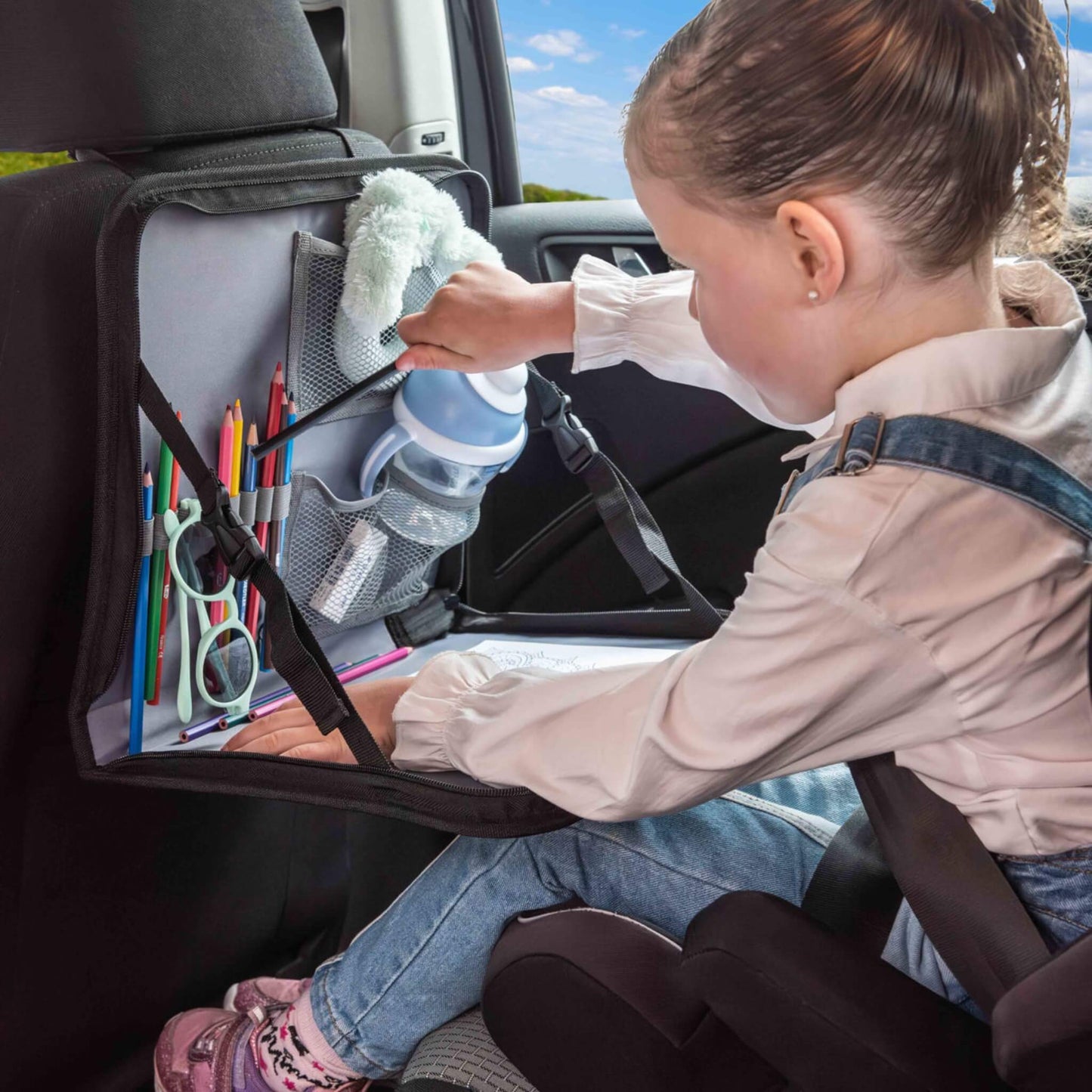 A young child seated in a car seat, picking out a pencil from inside the mesh pocket of a back seat travel tray filled with crayons and soft toys.