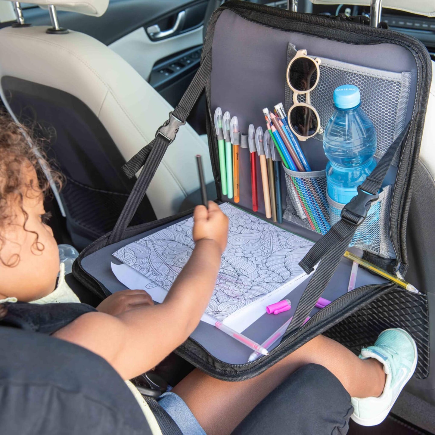 A toddler drawing on a butterfly colouring sheet, using a lap tray filled with colourful pens and a bottle secured in the mesh pocket.