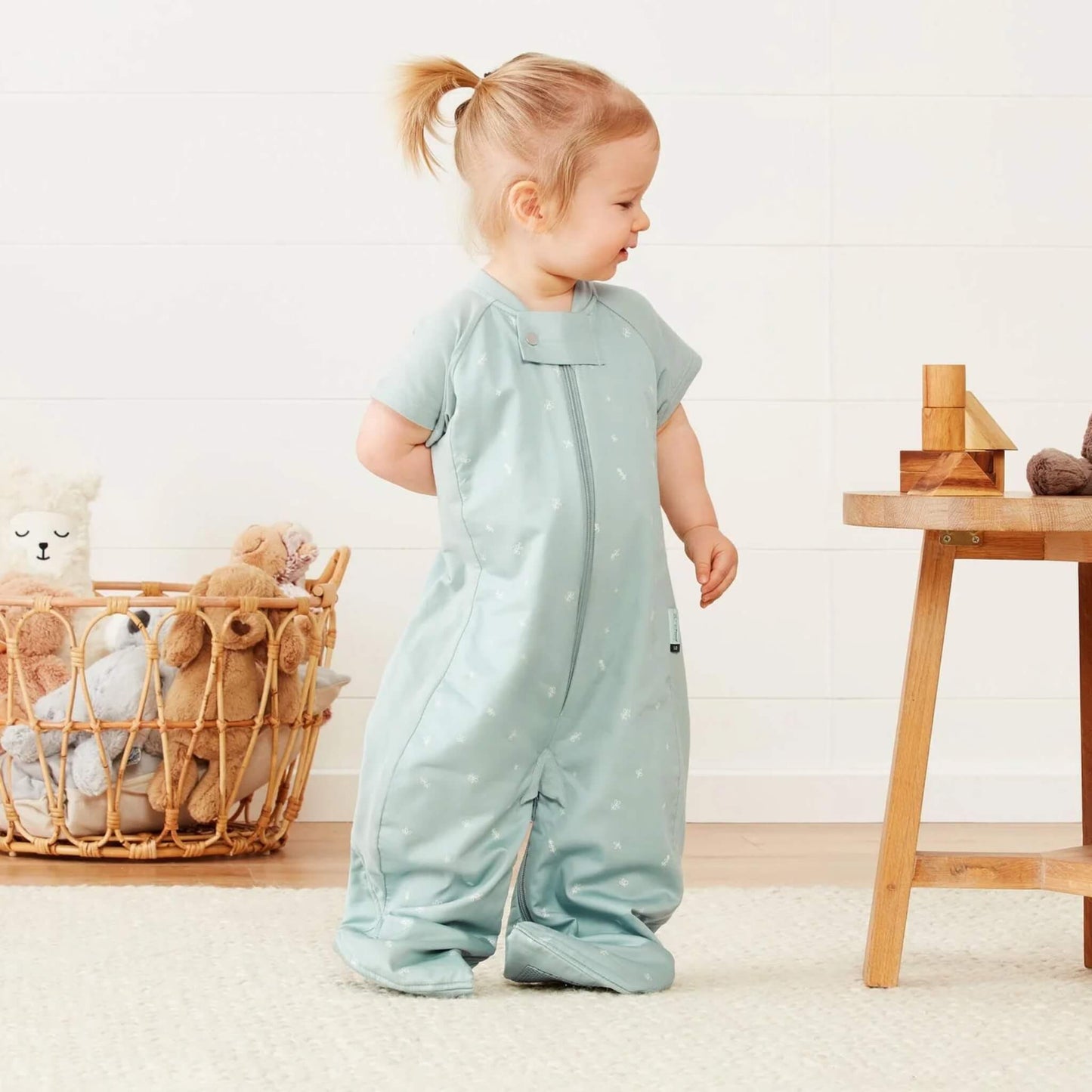 Toddler wearing a sage green short sleeved sleep suit bag in suit mode with separated legs, standing beside a wooden table and toys.
