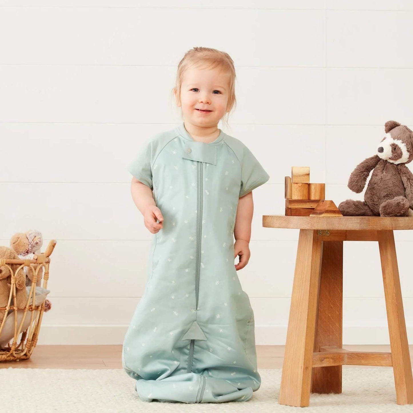 Toddler standing in a sage green short sleeved sleep suit bag, worn in sleeping bag style with legs enclosed.