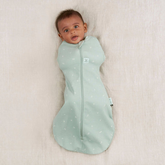 A baby comfortably swaddled in a Cocoon Swaddle Bag, featuring a soft sage green fabric with a secure zip-up front design.