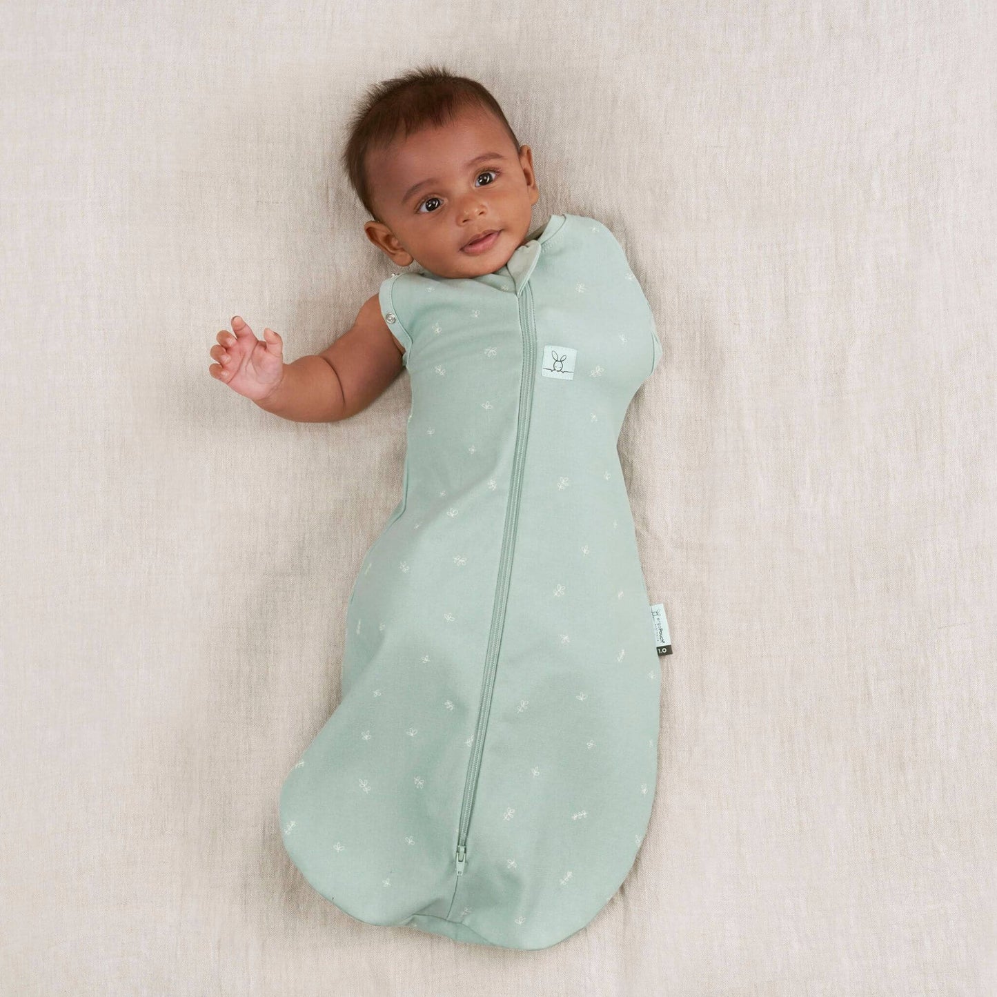 A baby lying on a soft beige surface, wearing a Swaddle Bag in sage green, transitioning with one arm out.