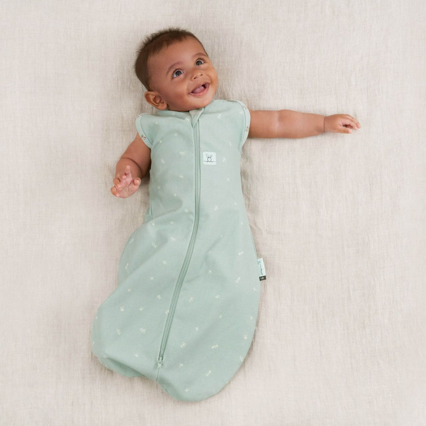A smiling baby wearing a Swaddle Bag in sage green, now in the arms-out transition mode with soft breathable fabric.
