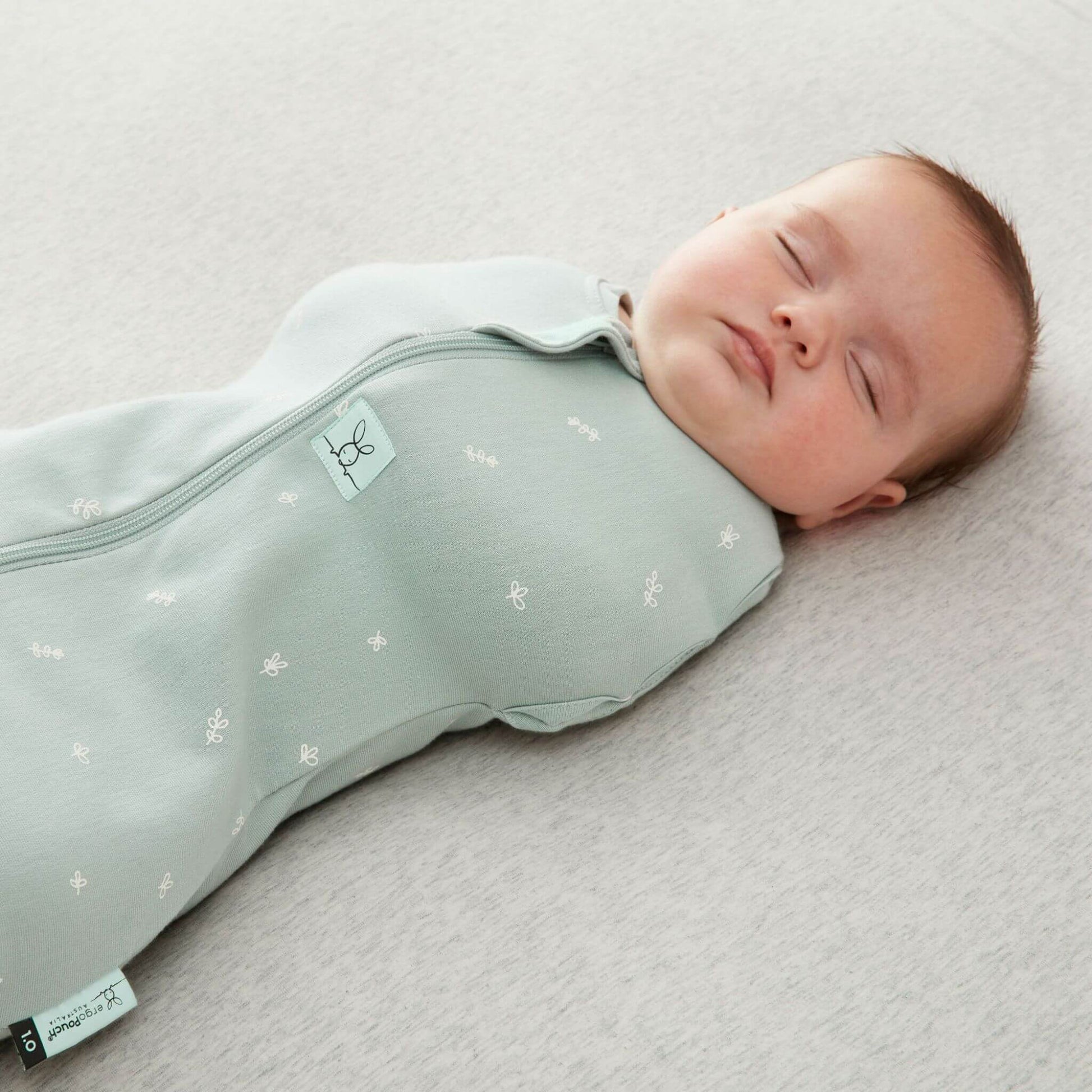 A peacefully sleeping baby in a  Swaddle Bag in sage green, lying on a soft grey surface.