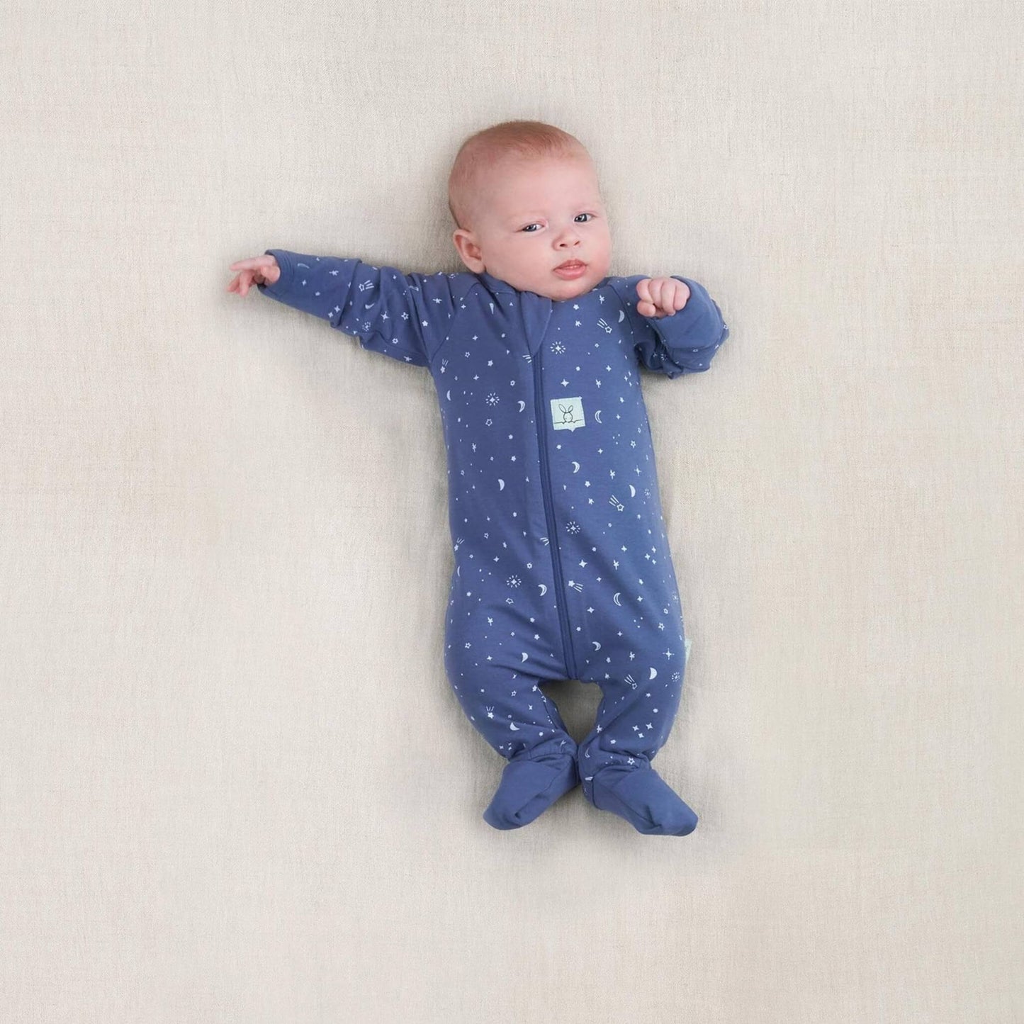 Newborn baby wearing a blue cotton babygrow with moon and star print, lying on a neutral surface with arms outstretched.
