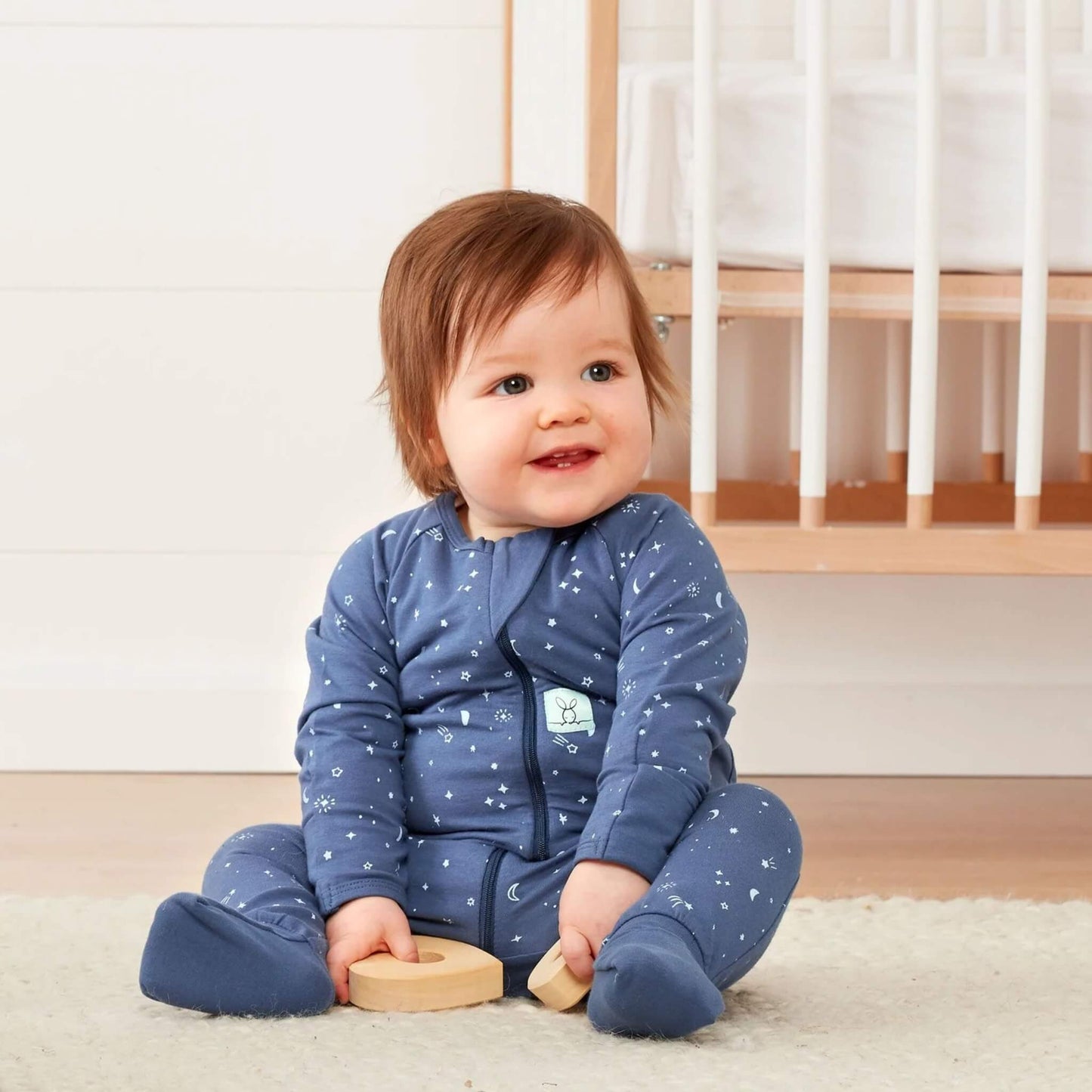 Smiling baby sitting on the floor wearing a blue starry night babygrow, holding wooden toys with a cot in the background.