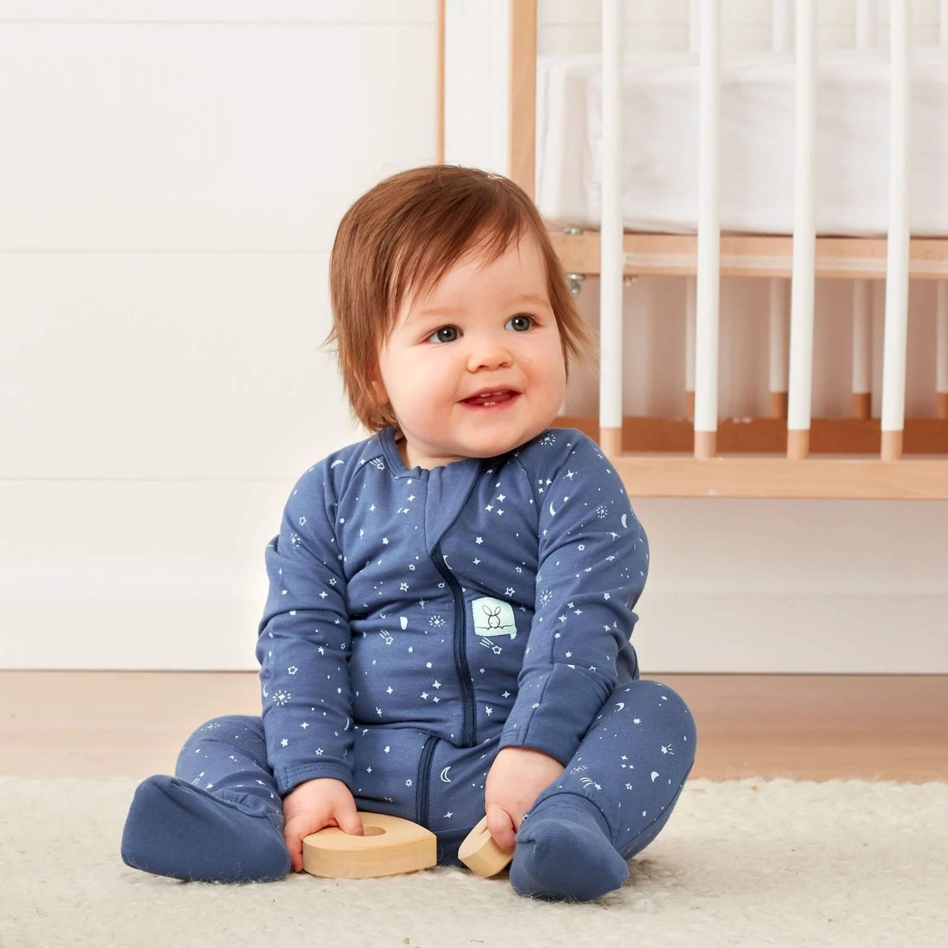 Smiling baby sitting on the floor wearing a blue starry night babygrow, holding wooden toys with a cot in the background.