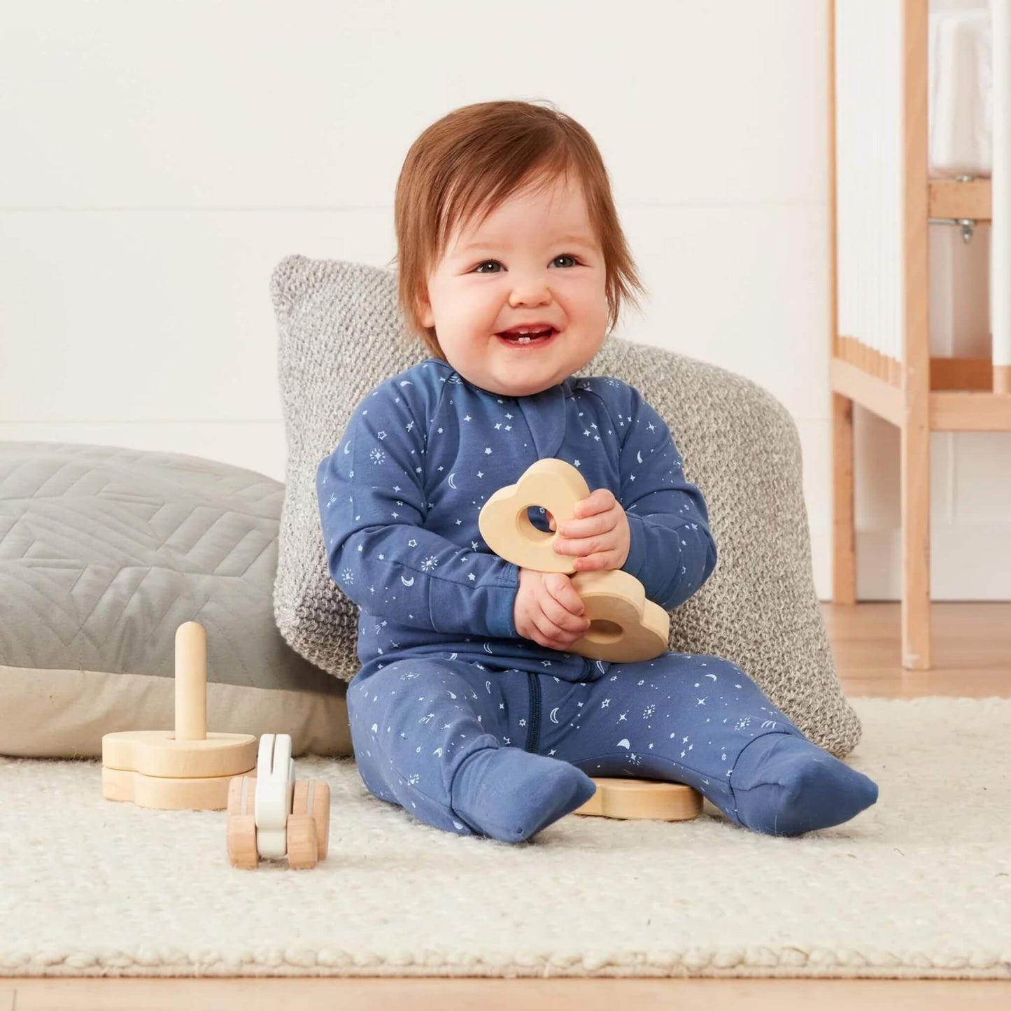 Baby in a moon and star print babygrow sitting on a soft rug with a cushion behind, holding a wooden toy and smiling.