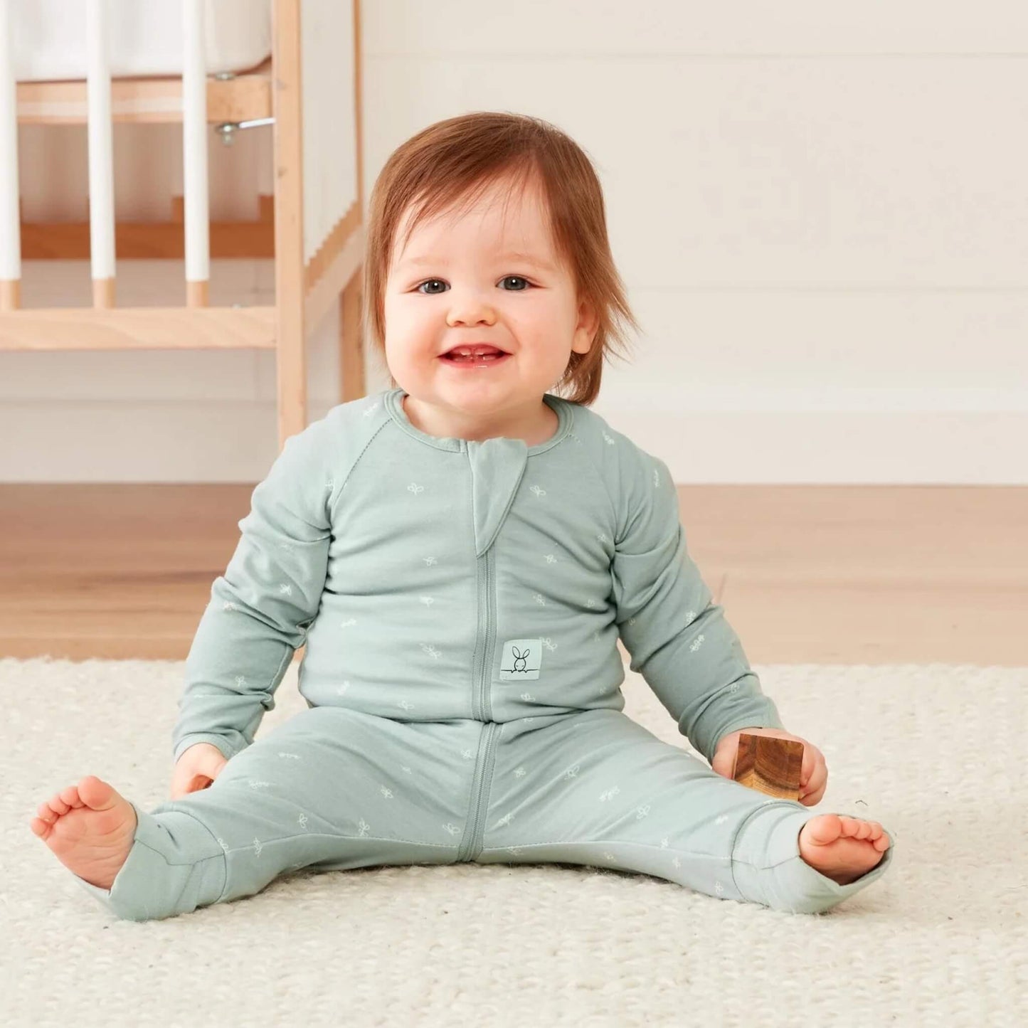 Smiling baby sitting on a rug in a Sage green cotton babygrow with zip front, holding a wooden block with a cot in the background.