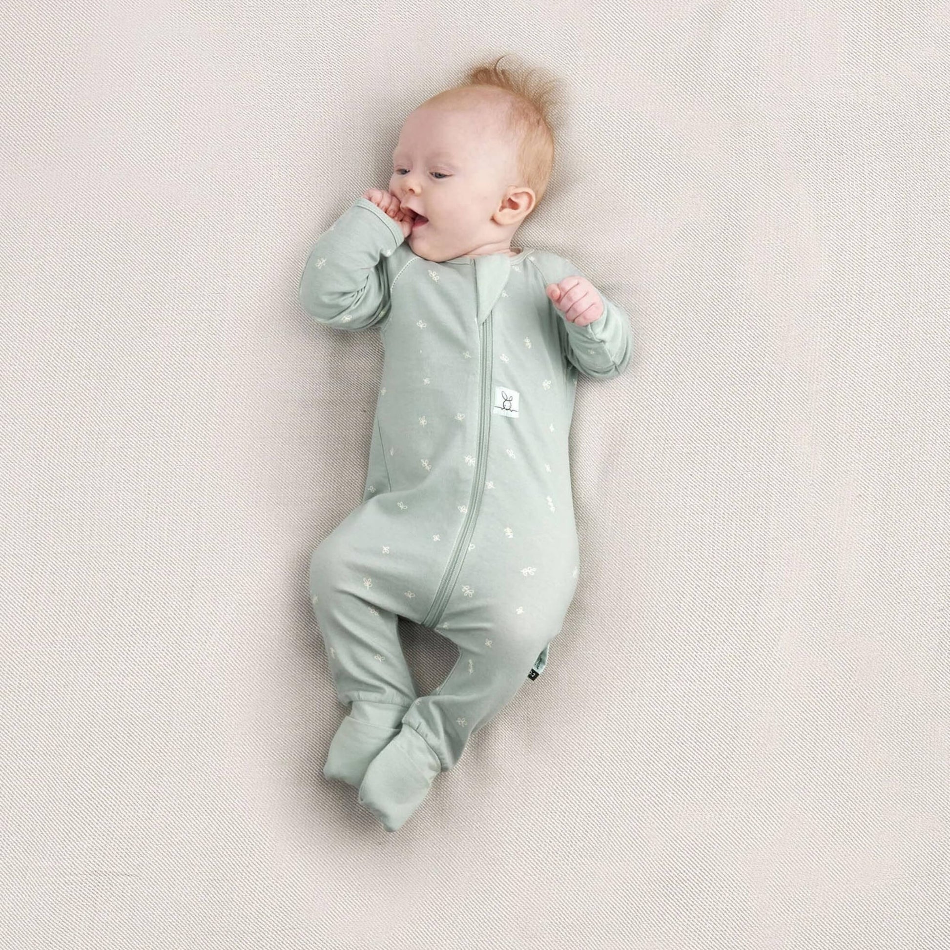 Baby lying on their side with thumb near mouth, dressed in a Sage green long sleeve babygrow with white leaf print and fold-over feet.
