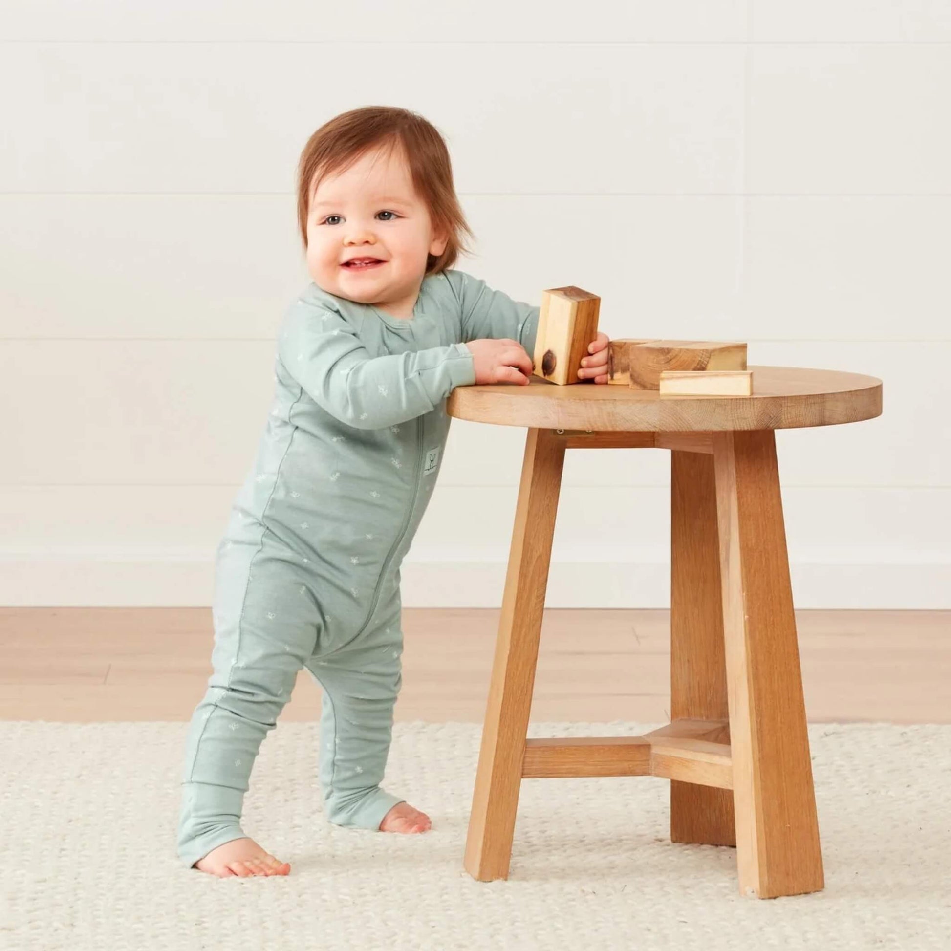 Baby in a Sage green long sleeve babygrow standing at a wooden stool, playing with blocks, with fold-over cuffs visible at the ankles.