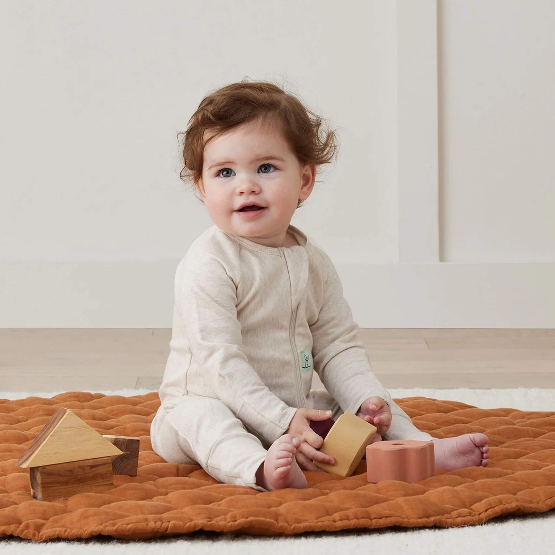 Baby sitting on a quilted rust-coloured mat, wearing a soft oatmeal babygrow and playing with wooden blocks.