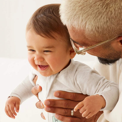 Smiling baby in an oatmeal long sleeve babygrow, being gently held by an adult with blond hair and glasses.