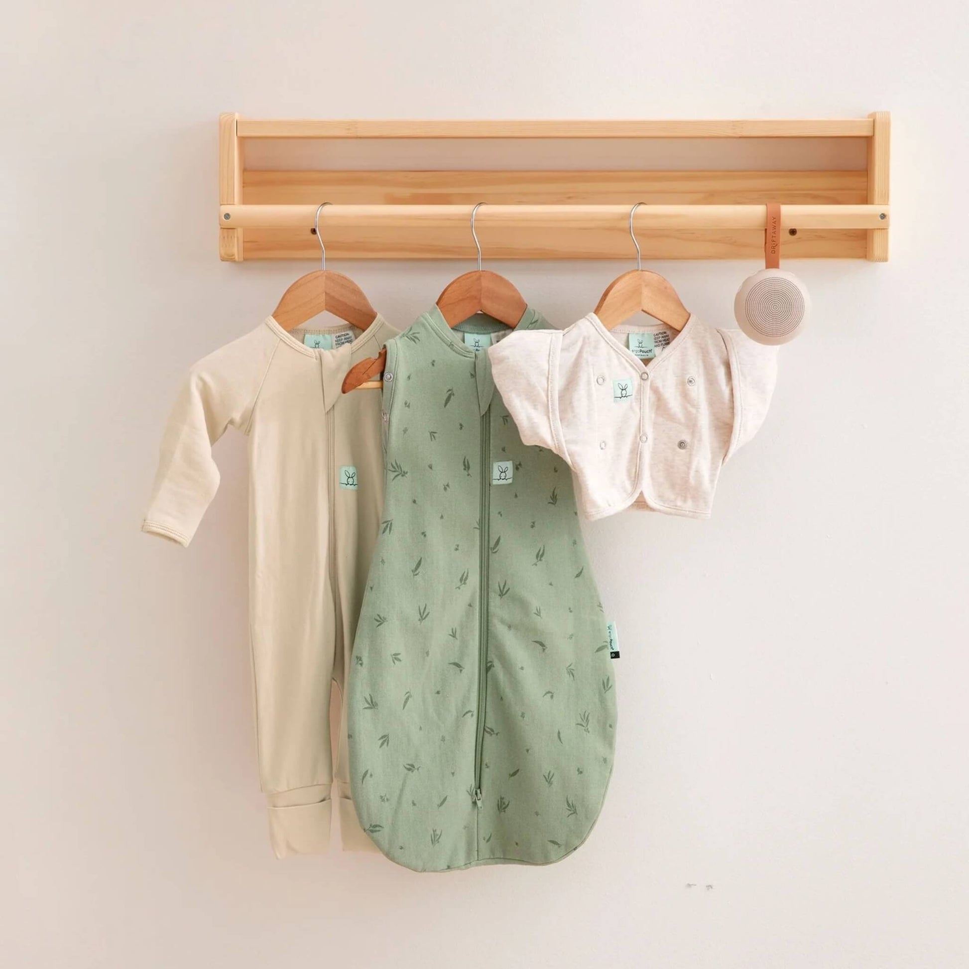 Three newborn sleepwear essentials hanging on wooden hangers, including a green swaddle bag, cream sleepsuit, and soft newborn cardi, with a portable sound machine hanging beside them.