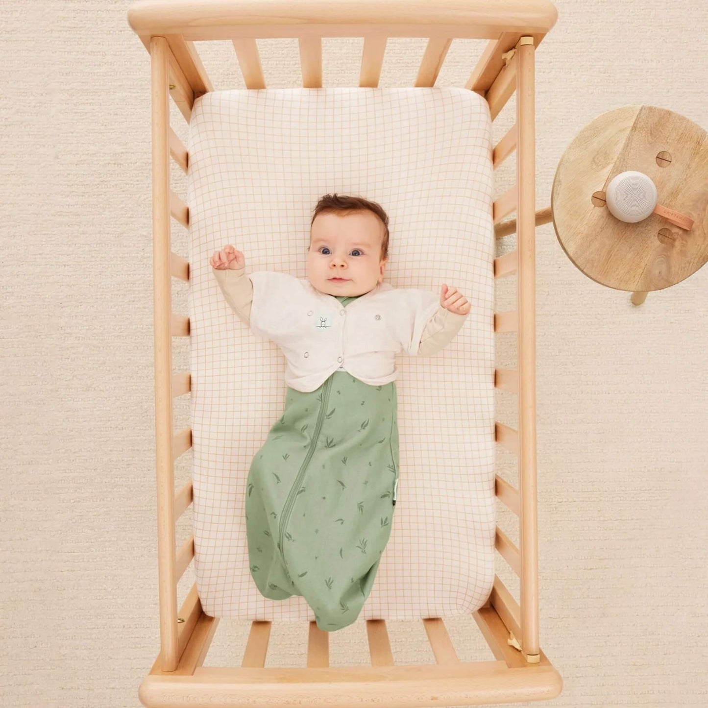 A baby lies in a wooden cot wearing a light cardigan and green patterned sleeping bag with arms tucked inside, resting on a cream and orange check mattress.