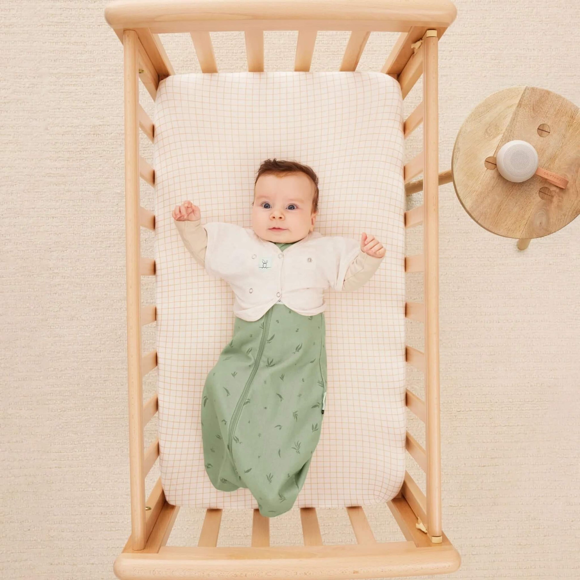 A baby lies in a wooden cot wearing a light cardigan and green patterned sleeping bag with arms tucked inside, resting on a cream and orange check mattress.