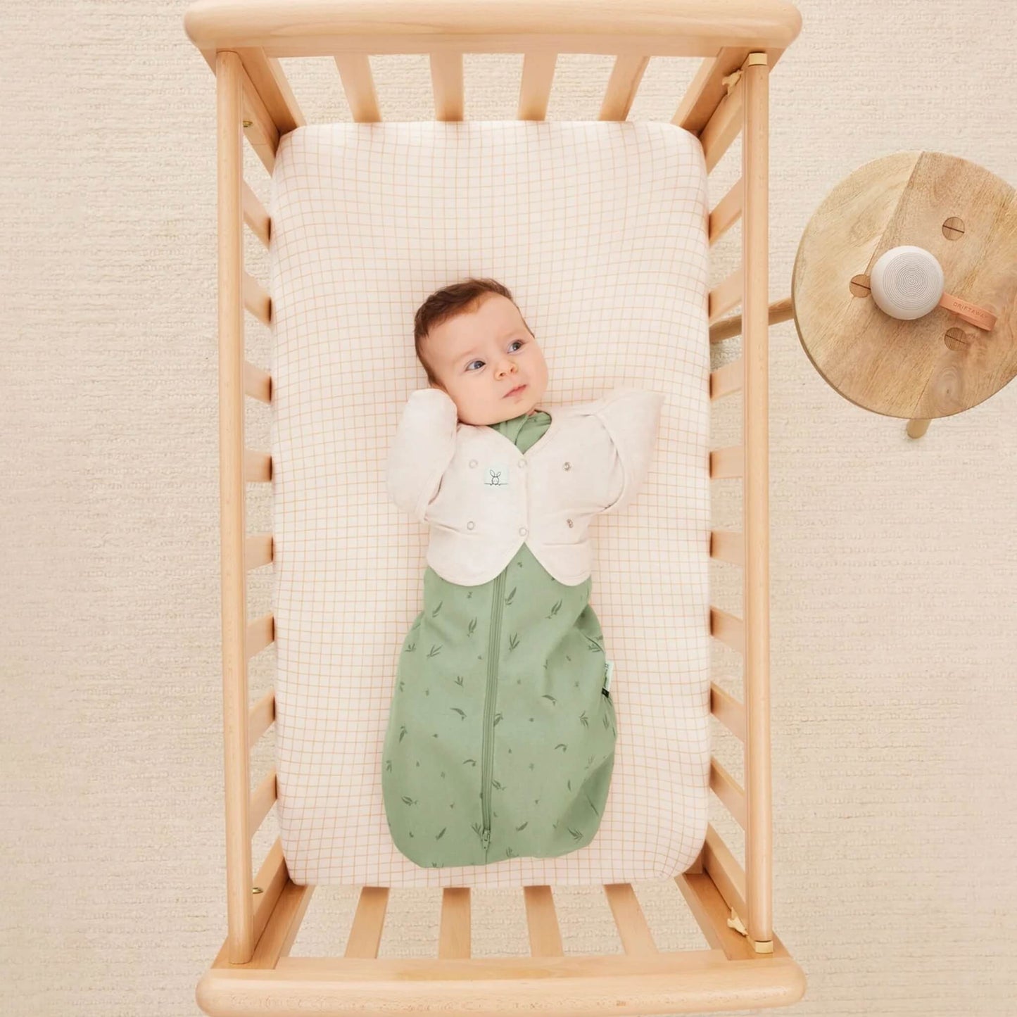 A baby lies in a wooden cot wearing a light cardigan and green patterned sleeping bag with arms stretched out, resting on a cream and orange check mattress.