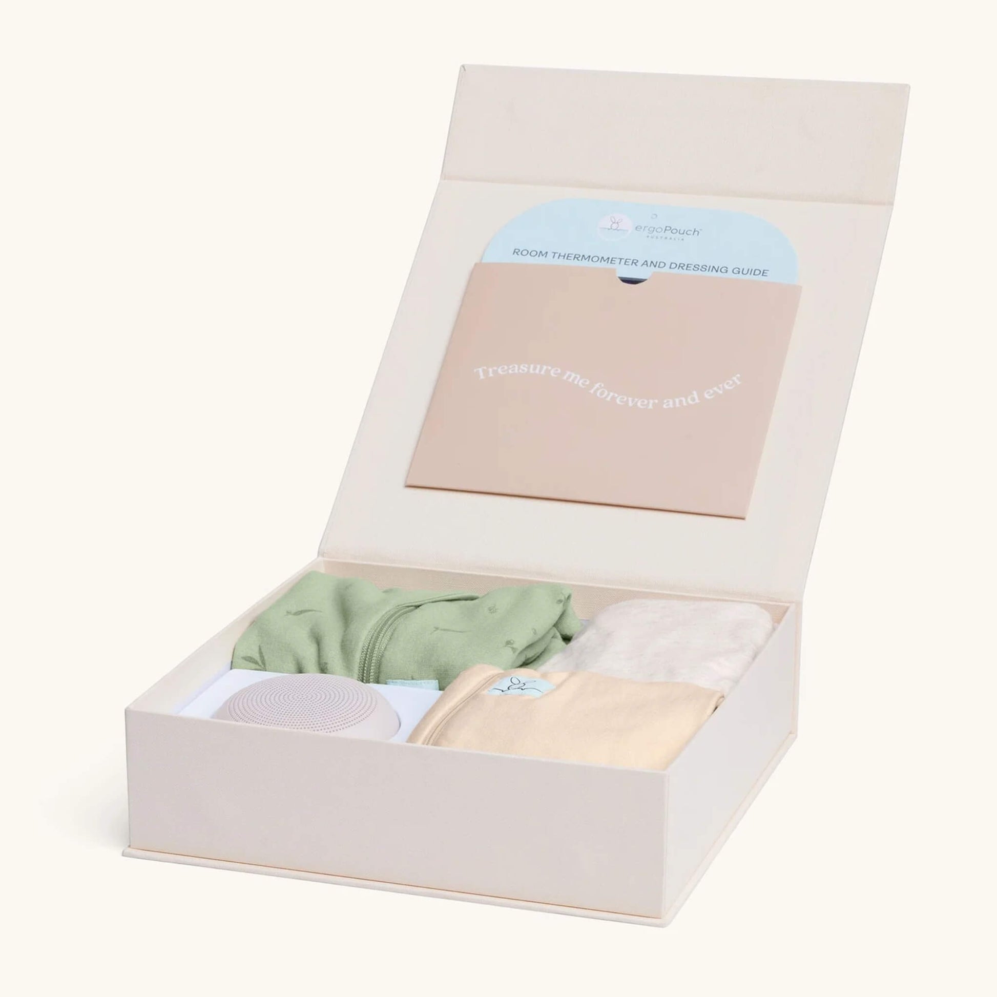An open keepsake box neatly holding newborn sleepwear and accessories, including a green swaddle bag, cream sleepsuit, cardi, and portable sound machine.