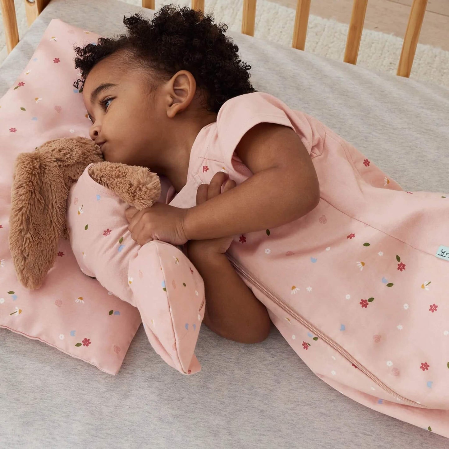 Toddler lying in a cot, cuddling a soft toy while resting on a pink floral pillow with matching bedding.