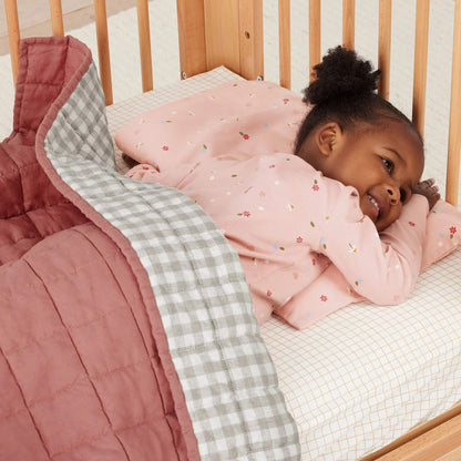Toddler lying under a quilt in a cot, smiling while resting on a pink pillow decorated with flowers, leaves, and ladybirds.