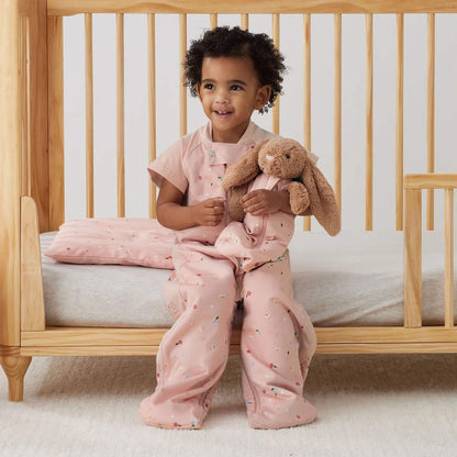 Toddler sitting on the edge of a wooden cot, holding a soft bunny toy with matching floral fabric, with the pillow placed behind.